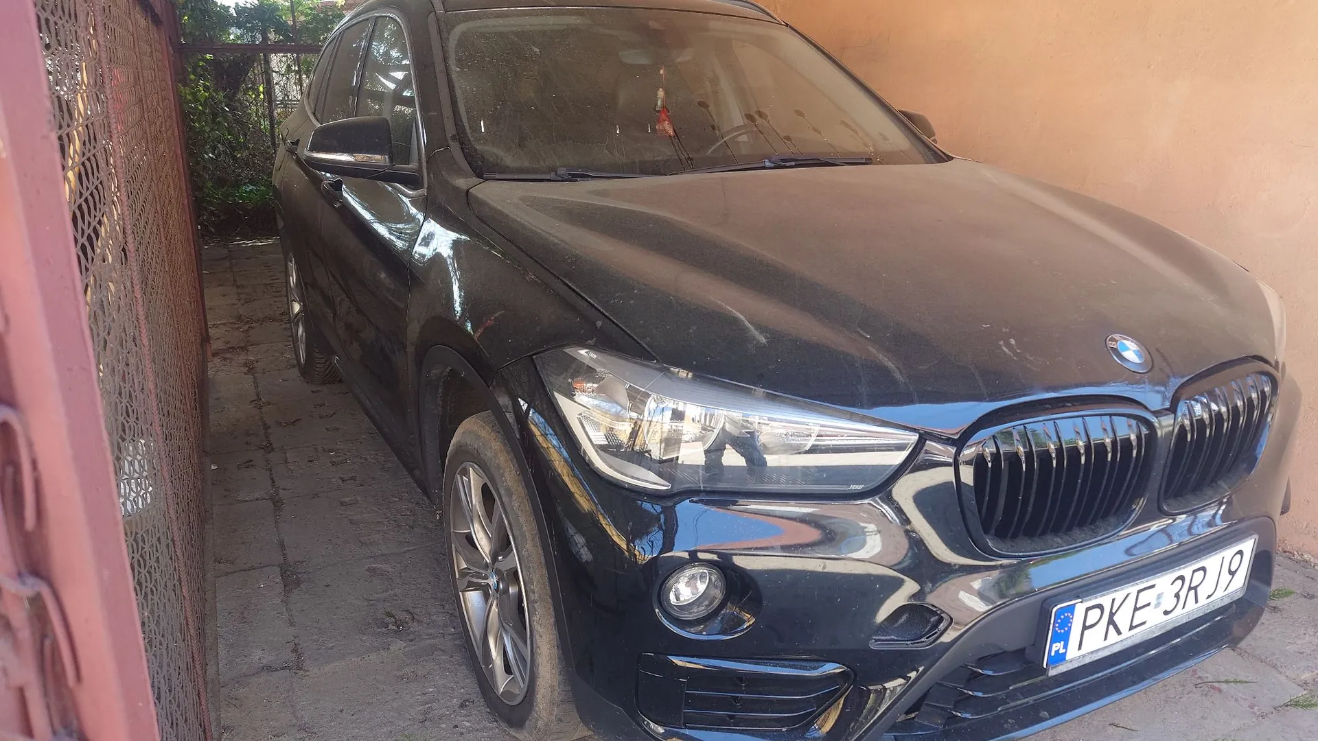 Czarny samochód marki BMW zaparkowany w wąskim przejeździe między różową bramą a ścianą budynku. Widoczny jest przód pojazdu z charakterystyczną atrapą chłodnicy i tablicą rejestracyjną o numerze PKE 3R19.