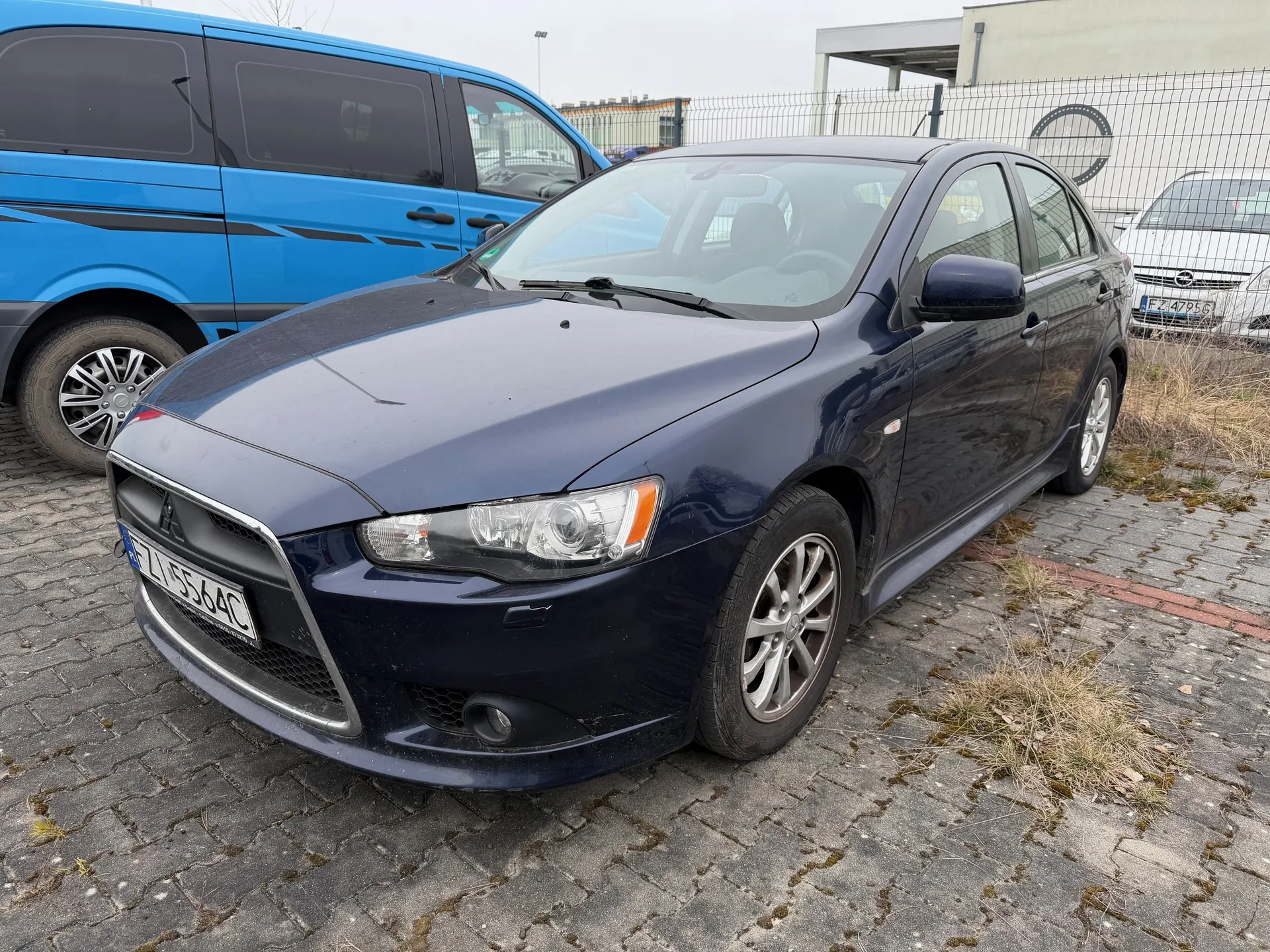 Ciemnoniebieski samochód osobowy marki Mitsubishi z tablicą rejestracyjną FZ 5564C zaparkowany na brukowanym placu. W tle niebieski van z czarnymi pasami, biały samochód z tablicą FZ 4795 oraz metalowe ogrodzenie. Na pierwszym planie widoczne suchą trawę i fragment budynku.