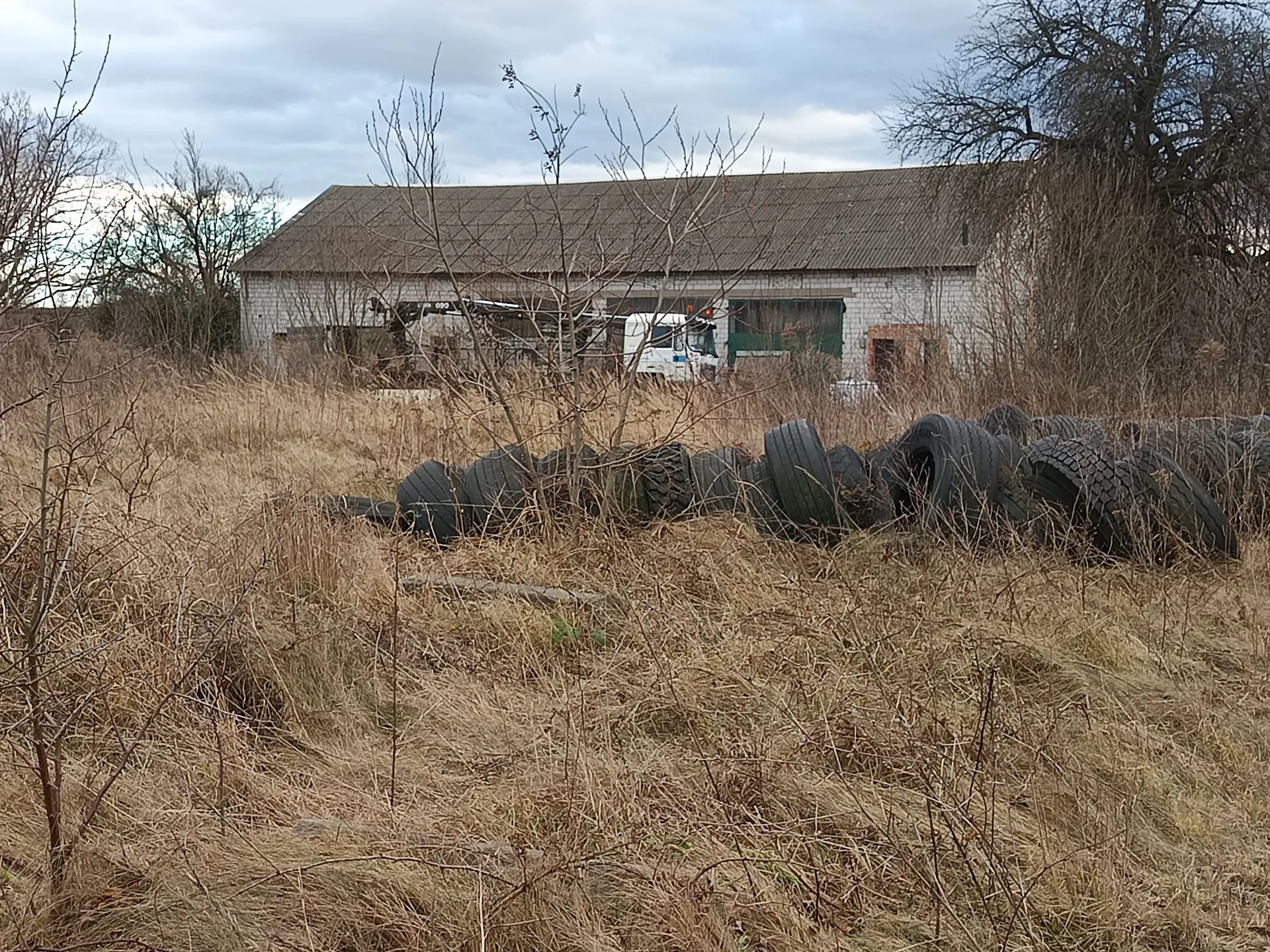 Krajobraz z jesienną lub zimową roślinnością w kolorach brązu i beżu. Na pierwszym planie znajduje się wyschnięta trawa i krzewy. W środkowej części obrazu widoczny jest rząd zużytych opon samochodowych ułożonych w linii. W tle znajduje się długi, niski budynek gospodarczy z szarym dachem i białymi ścianami, przed którym zaparkowany jest biały samochód dostawczy. Nad budynkiem widać zachmurzone niebo.