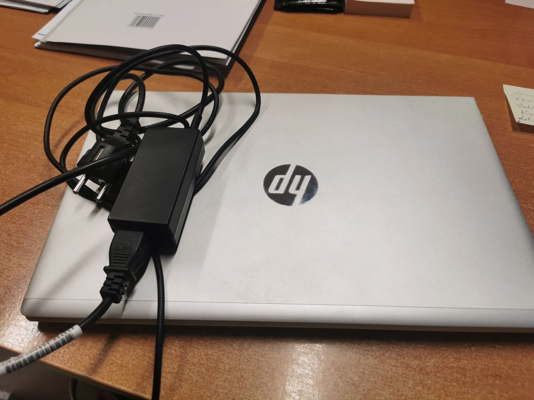 Srebrny laptop marki HP zamknięty i leżący na drewnianym stole. Na górnej obudowie laptopa znajduje się czarny zasilacz z zaplątanym kablem zasilającym.