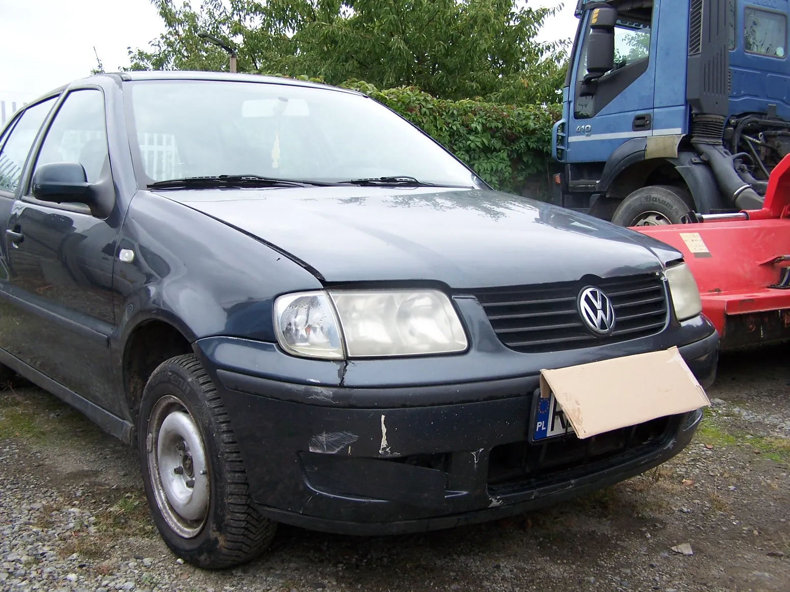 Ciemnogranatowy samochód osobowy marki Volkswagen zaparkowany na żwirowym podłożu. Auto posiada widoczne uszkodzenia z przodu, w tym zarysowania i wgniecenia na zderzaku. Tablica rejestracyjna jest częściowo zasłonięta kawałkiem brązowej tektury. W tle widoczny jest niebieski samochód ciężarowy oraz zielone krzewy i drzewa.