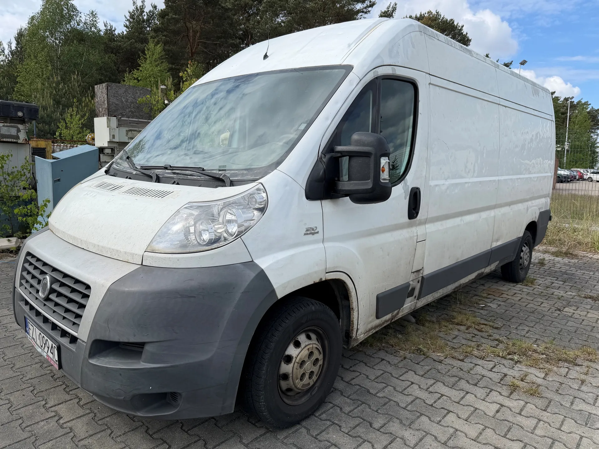 Biały furgon marki Fiat z widocznym logo na masce, z tablicą rejestracyjną ZL 0994C. Pojazd ma szare elementy na przednim zderzaku i nadkółach, z widocznymi śladami brudu. W tle drzewa, przemysłowe konstrukcje, ogrodzenie i inne samochody na parkingu. Niebo częściowo zachmurzone. Furgon stoi na brukowanej powierzchni z miejscami porośniętymi trawą.