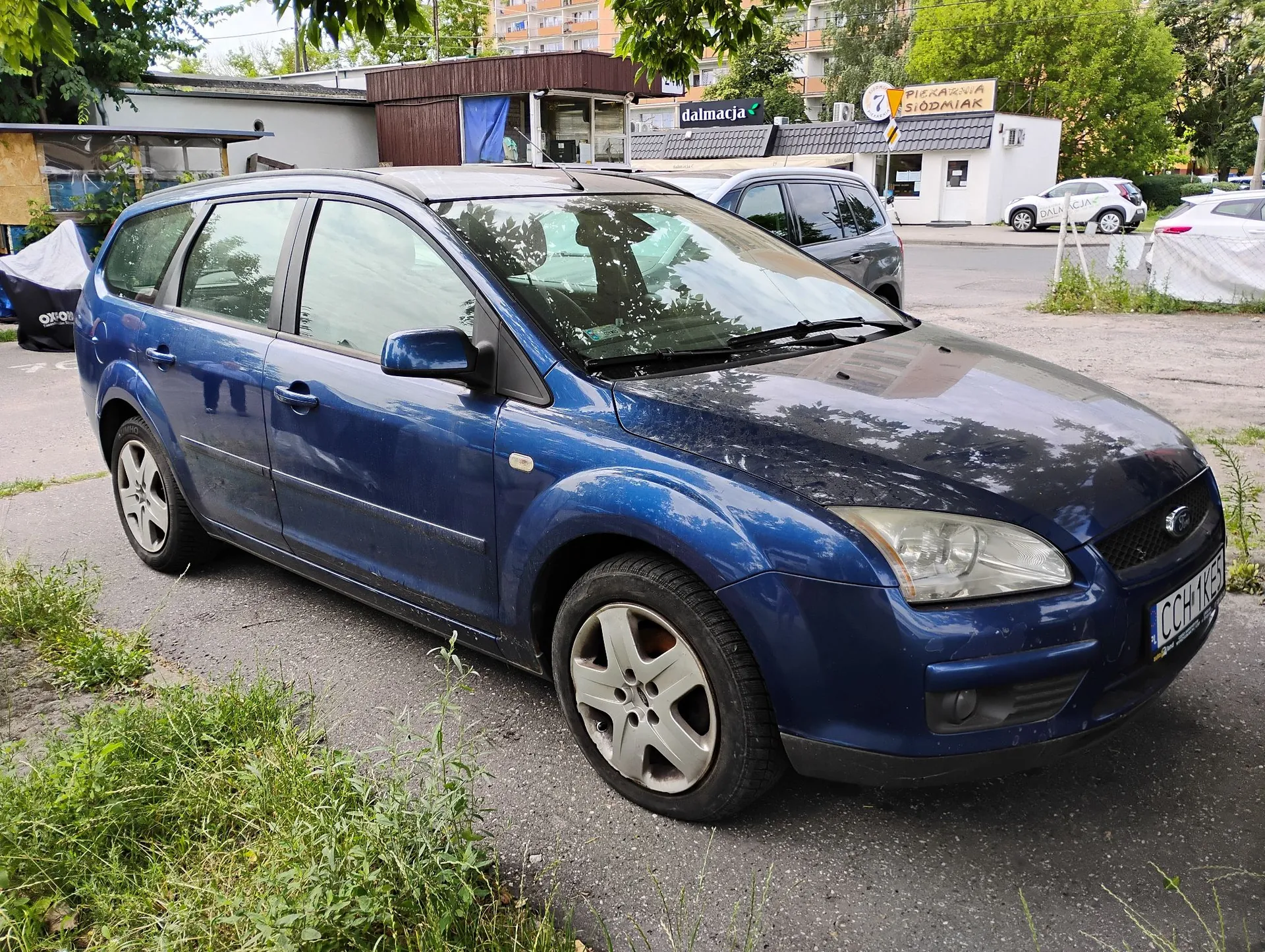 Niebieski samochód typu kombi zaparkowany na asfaltowej drodze. W tle widoczne są budynki handlowe z napisami „dalmacja” oraz „PIEKARNIA SIÓDMIAK”, a także inne zaparkowane samochody i zieleń miejska.