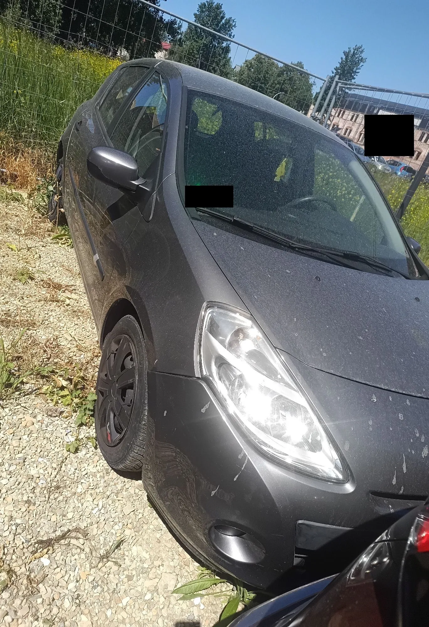 Zdjęcie przedstawiające przód i bok czarnego samochodu osobowego zaparkowanego na żwirowym podłożu. Pojazd jest zakurzony. W tle widoczny jest metalowy płot z siatką, zieleń oraz fragmenty innych budynków i samochodów pod jasnym, niebieskim niebem.