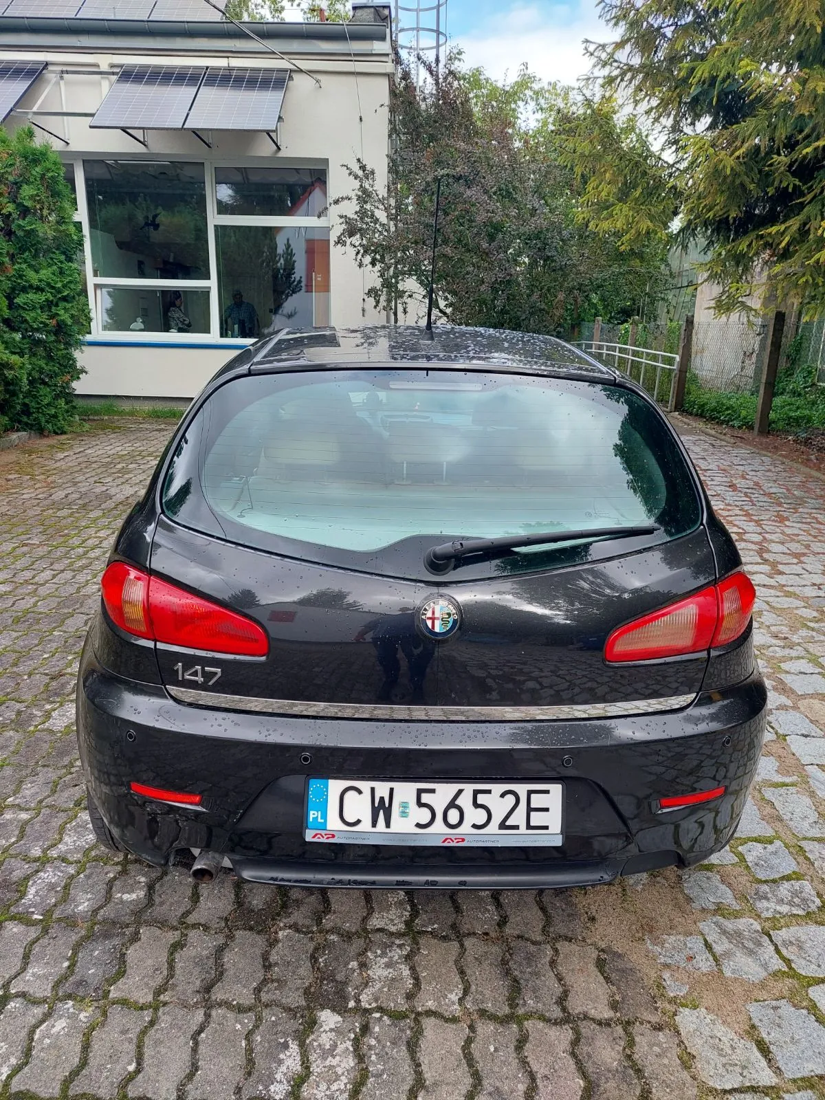 Tył czarnego samochodu marki Alfa Romeo model 147 zaparkowanego na kostce brukowej. Samochód ma polską tablicę rejestracyjną o numerze CW 5652E. W tle widoczny jest jasny budynek z panelami fotowoltaicznymi na dachu oraz zielone drzewa i krzewy.