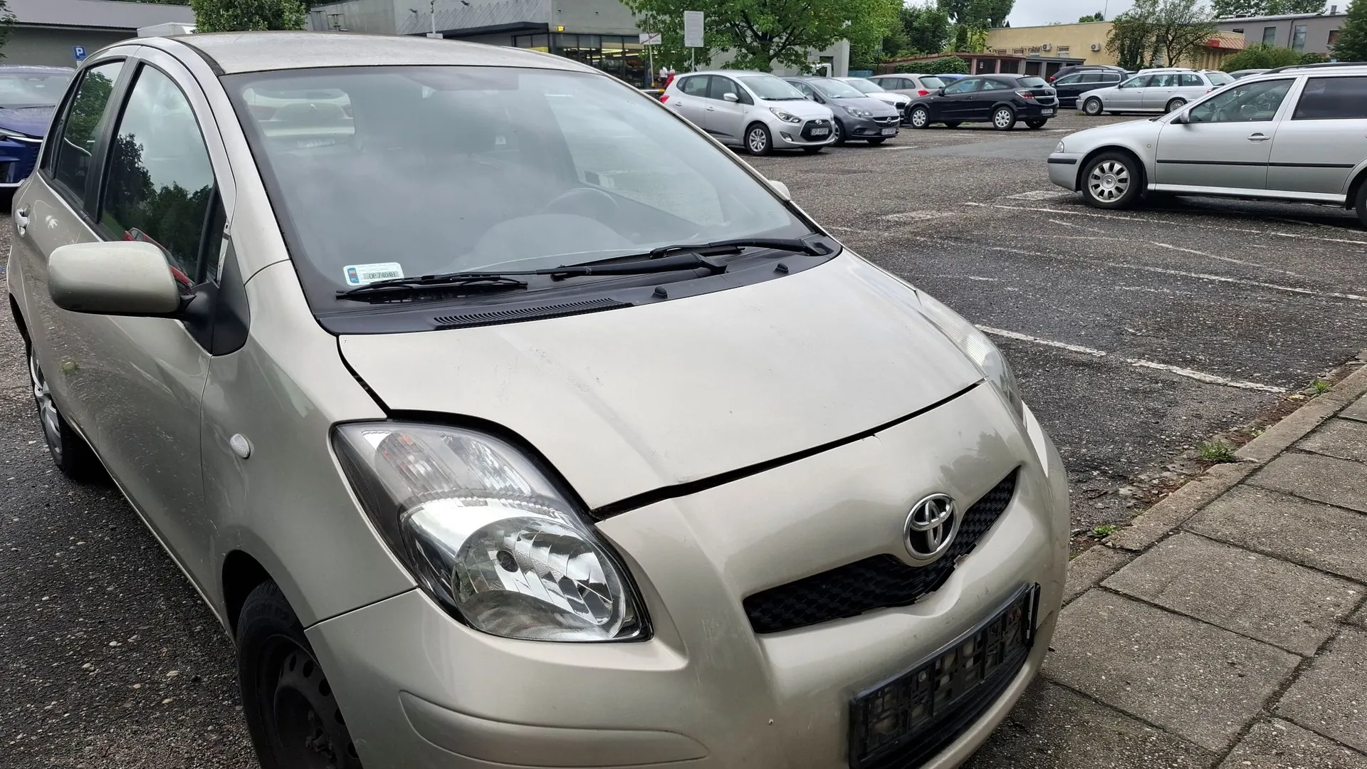Jasnoszary samochód osobowy marki Toyota zaparkowany na asfaltowym parkingu. Pojazd jest widoczny z przodu i z boku, z lekko uchyloną maską silnika. W tle widoczne są inne zaparkowane samochody oraz budynki i drzewa.