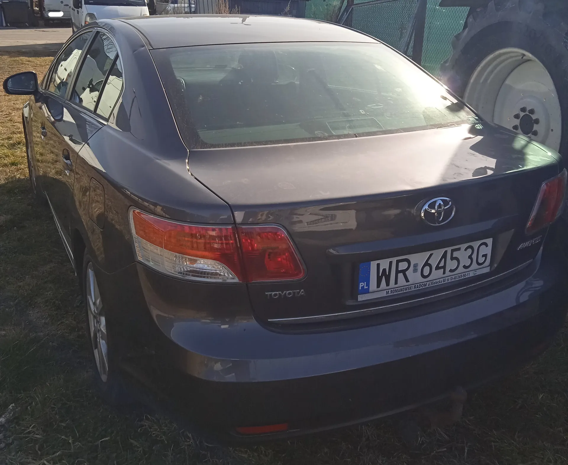 Ciemnoszary samochód osobowy marki Toyota, widok od tyłu. Na bagażniku widoczne logo Toyota oraz tablica rejestracyjna z napisem PL WR 6453G. Po prawej stronie obrazu duży opona ciągnika, w tle ogrodzenie i inne pojazdy. Samochód stoi na trawie, słońce oświetla tylną część pojazdu.