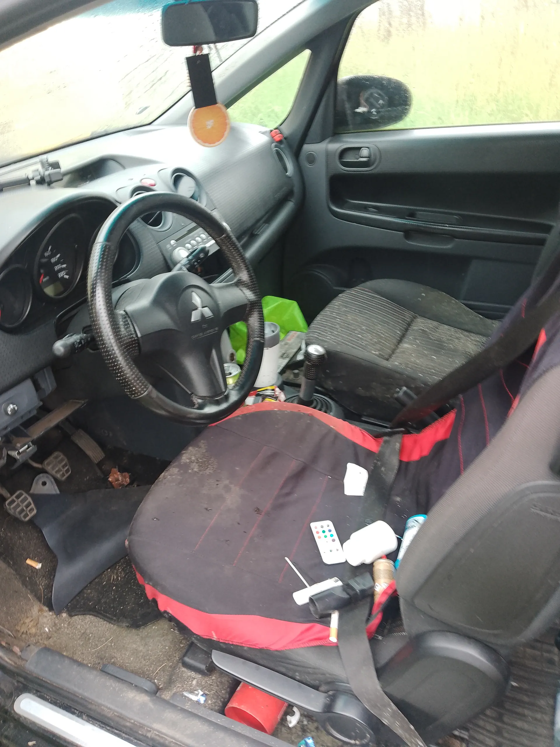 Wnętrze samochodu z widocznym kierownicą marki Mitsubishi, zniszczonymi siedzeniami z czarno-czerwonymi pokrowcami, rozrzuconymi przedmiotami (pilot, papieros, narzędzia, puszka), zanieczyszczonym podłogą i deską rozdzielczą. W tle przez otwarte drzwi widać trawę. Na lusterku wstecznym wiszący pomarańczowy odświeżacz powietrza.
