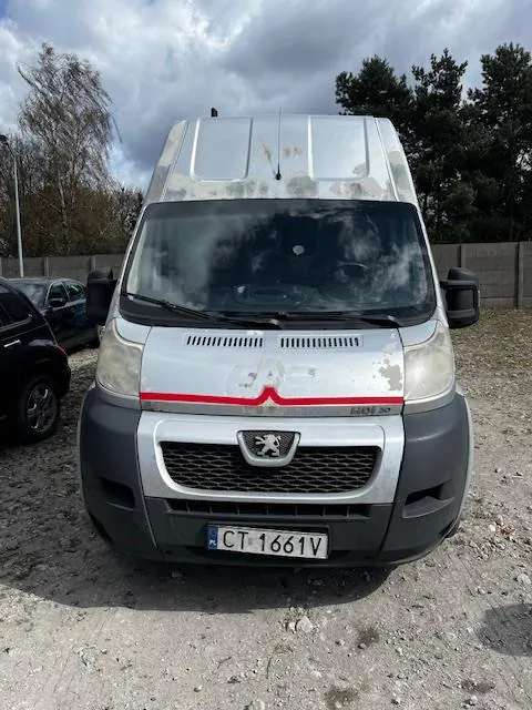 Przedni widok ciężarówki marki Peugeot, białej z szarymi elementami i czerwoną linią na masce. Na masce widoczny jest logo Peugeota oraz tablica rejestracyjna CT-1661V. Po lewej stronie częściowo widoczny czarny samochód. Tło stanowią drzewa, betonowy płot i zachmurzone niebo. Podłoga z żwiru. Na masce widoczne ślady zabrudzeń.