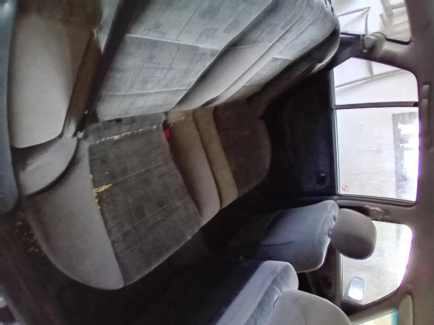 Wnętrze samochodu z widokiem na tylne siedzenia. Siedzenia są szare, zużyte, z widocznymi plamami i uszkodzeniami. Na środkowym siedzeniu znajduje się czerwony element, prawdopodobnie zamek pasów bezpieczeństwa. Widoczne są przednie siedzenia z zagłówkami oraz okna boczne i tylne. Za oknami widać fragment budynku i zewnętrzne otoczenie. Wnętrze wydaje się brudne, a podłoga pokryta jest śmieciem.