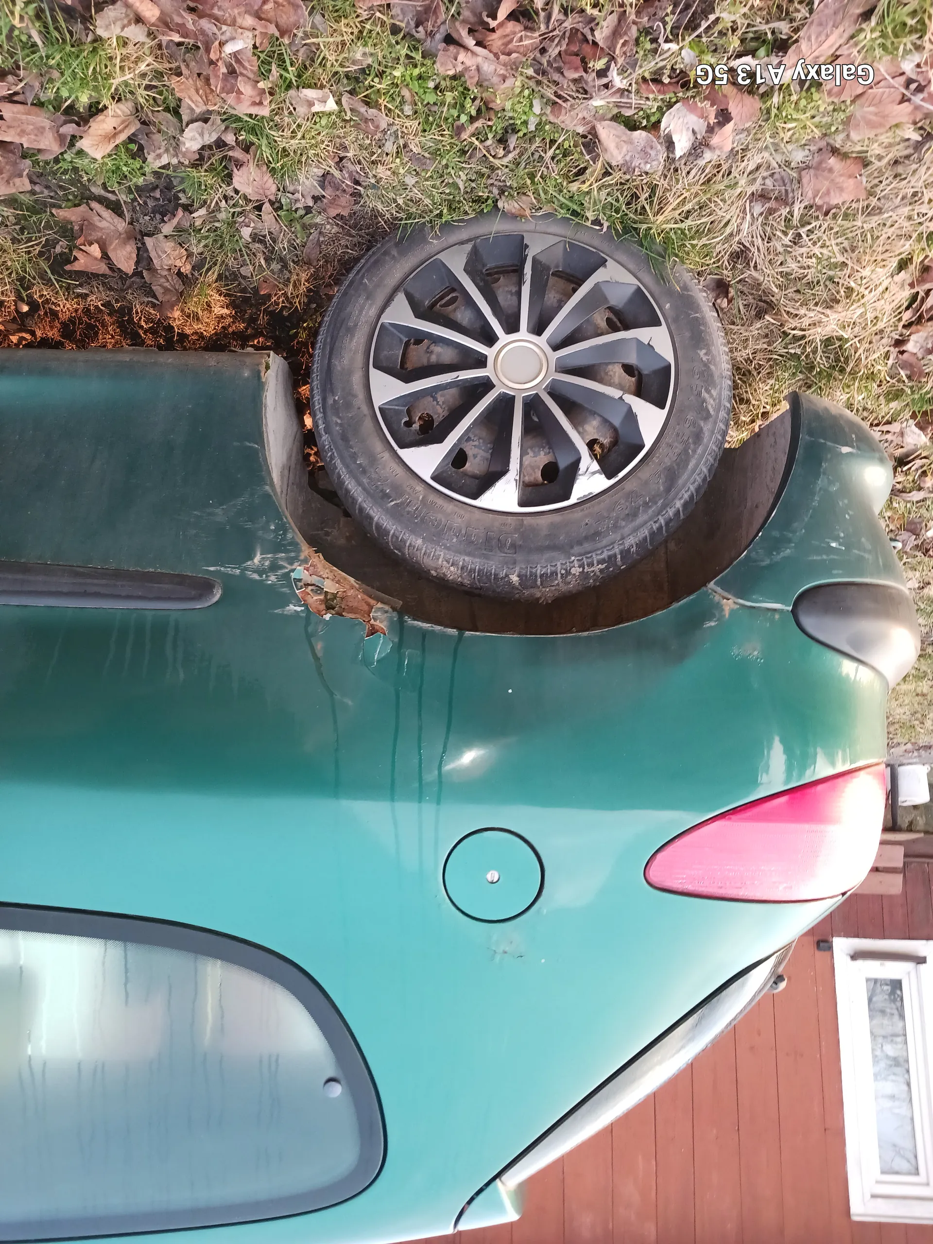 Zielony samochód z widocznym czerwonym światłem tylnym, kołem z felgą w kształcie gwiazdy oraz rdzą wokół koła. W tle trawa z suchymi liśćmi i drewniana konstrukcja z białym oknem. Po prawej stronie obrazu widoczny tekst: Galaxy A13 5G.