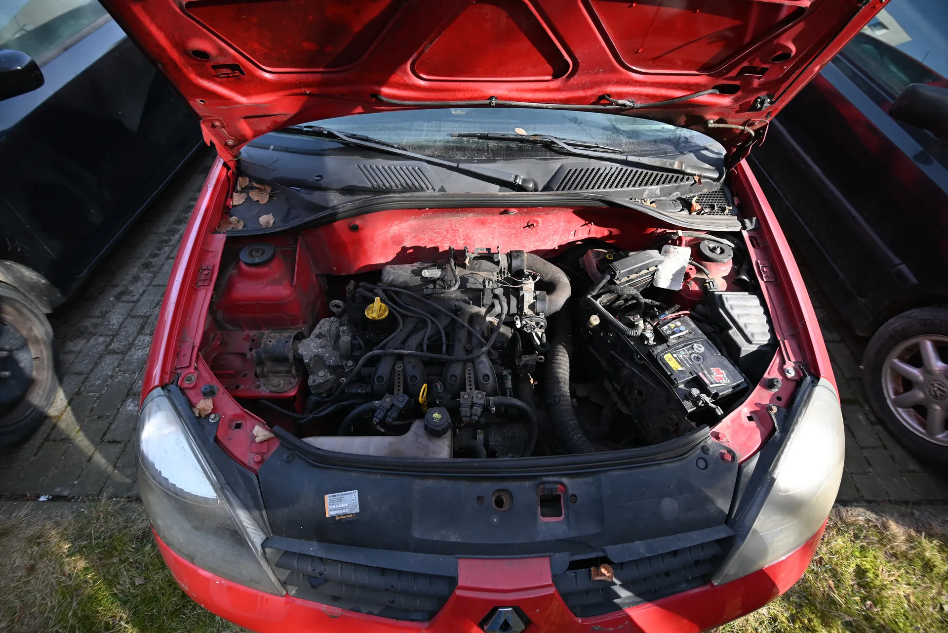 Czerwony samochód z otwartą maską ukazujący komorę silnika z widocznymi elementami: akumulatorem, żółtą nakrętką, przewodami i innymi częściami mechanicznymi. W komorze silnika znajdują się suche liście. Po obu stronach czerwonego samochodu widoczne są czarne pojazdy. Tło stanowi brukowana powierzchnia i trawa. Na przedniej części komory silnika znajduje się mała naklejka z nieczytelnym tekstem.