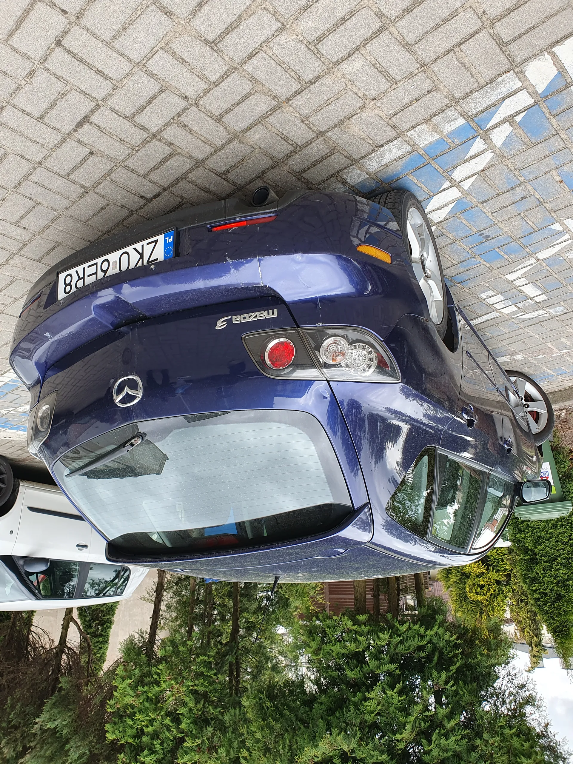 Tylne widok niebieskiego samochodu Mazda 3 zaparkowanego na brukowanej powierzchni. Widoczna tablica rejestracyjna ZK06ER8 z niebieskim pasem UE i oznaczeniem PL. Obok niebieskiego samochodu stoi biały samochód. W tle zielone krzewy i drzewa oraz fragment budynku.