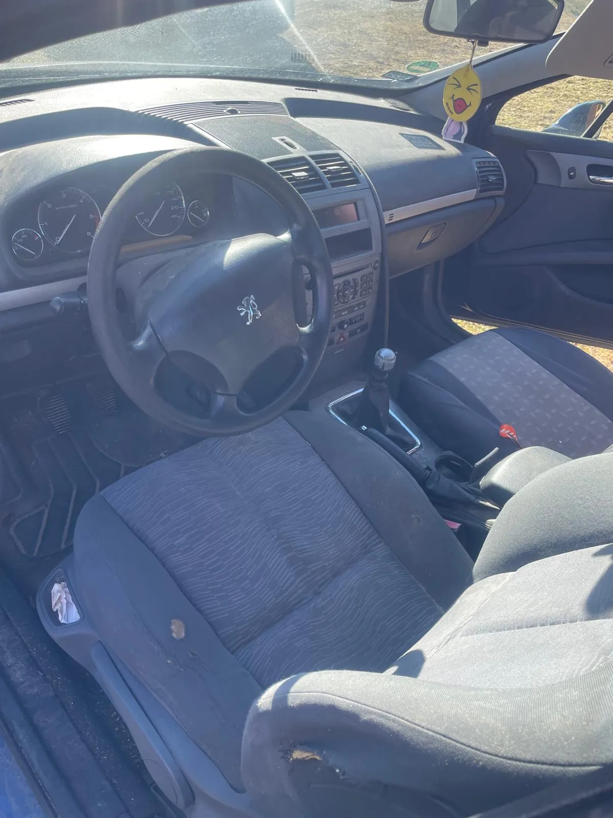 Wnętrze samochodu marki Peugeot z widocznym kierownicą z logo, deska rozdzielcza z licznikami, centralną konsolą i dźwignią zmiany biegów. Siedzenia tapicerowane szarym materiałem z widocznymi uszkodzeniami i plamami. W lusterku wstecznym wisząca żółta ozdoba w kształcie emotikonki z otwartymi ustami. Drzwi pasażera są otwarte, za nimi widoczny suchy trawnik. Wnętrze oświetlone światłem słonecznym.