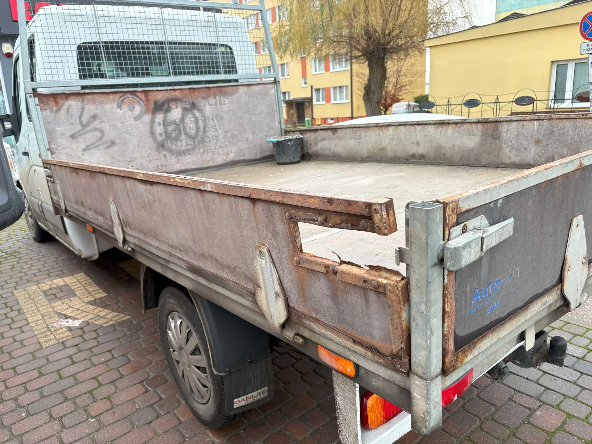 Zdjęcie przedstawia tył starszego, rdzewiejącego samochodu dostawczego z otwartym ladem. Na bocznej ścianie ladu widoczny jest napis: BORISOL, ISOLERING, STILLADS, INTERIOR, FACADE. Po prawej stronie ladu znajduje się niebieski napis Auto. W ladzie stoi czarny wiadro. Samochód stoi na brukowanej powierzchni, w tle widoczne są żółte budynki z czerwonymi oknami, drzewo oraz znak drogowy zakazu postoju. Opona tylna ma widoczną etykietę DANLAR. Konstrukcja ladu i elementy metalowe pokryte są rdzą.