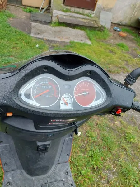 Zdjęcie przedstawiające kokpit czarnego skutera z widocznymi dwoma analogowymi wskaźnikami: górnym, mniejszym wskaźnikiem paliwa i temperatury oraz dolnym, większym prędkościomierzem z czerwoną wskazówką. Obraz jest zorientowany pionowo, podczas gdy skuter i tło (trawa, chodnik) znajdują się w pozycji poziomej.