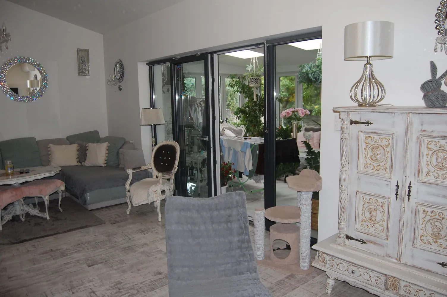 Jasny salon urządzony w stylu vintage i romantycznym. Po lewej stronie znajduje się szara narożna kanapa z jasnymi poduszkami, przed nią biały stolik kawowy i różowy taboret. W centrum stoi biały, ozdobny fotel w stylu klasycznym oraz szary fotel widziany od tyłu. Po prawej stronie znajduje się duża, biała, rzeźbiona szafka z lampką nocną na blacie. W tle duże przeszklone drzwi tarasowe prowadzące do ogrodu z roślinnością. Na ścianie wisi okrągłe lustro w ozdobnej ramie.