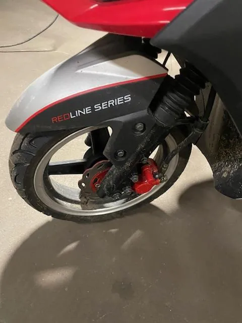 Zbliżenie przedniego koła motocykla z widocznym czerwonym i srebrnym nadkolem. Na czarnej części nadkola znajduje się napis 'REDLINE SERIES', gdzie 'REDLINE' jest czerwony, a 'SERIES' biały. Widoczne są również czarna opona z metalowym felgiem, czerwony zacisk hamulcowy oraz czarna amortyzacja. Tło stanowi betonowa podłoga z cieniami.