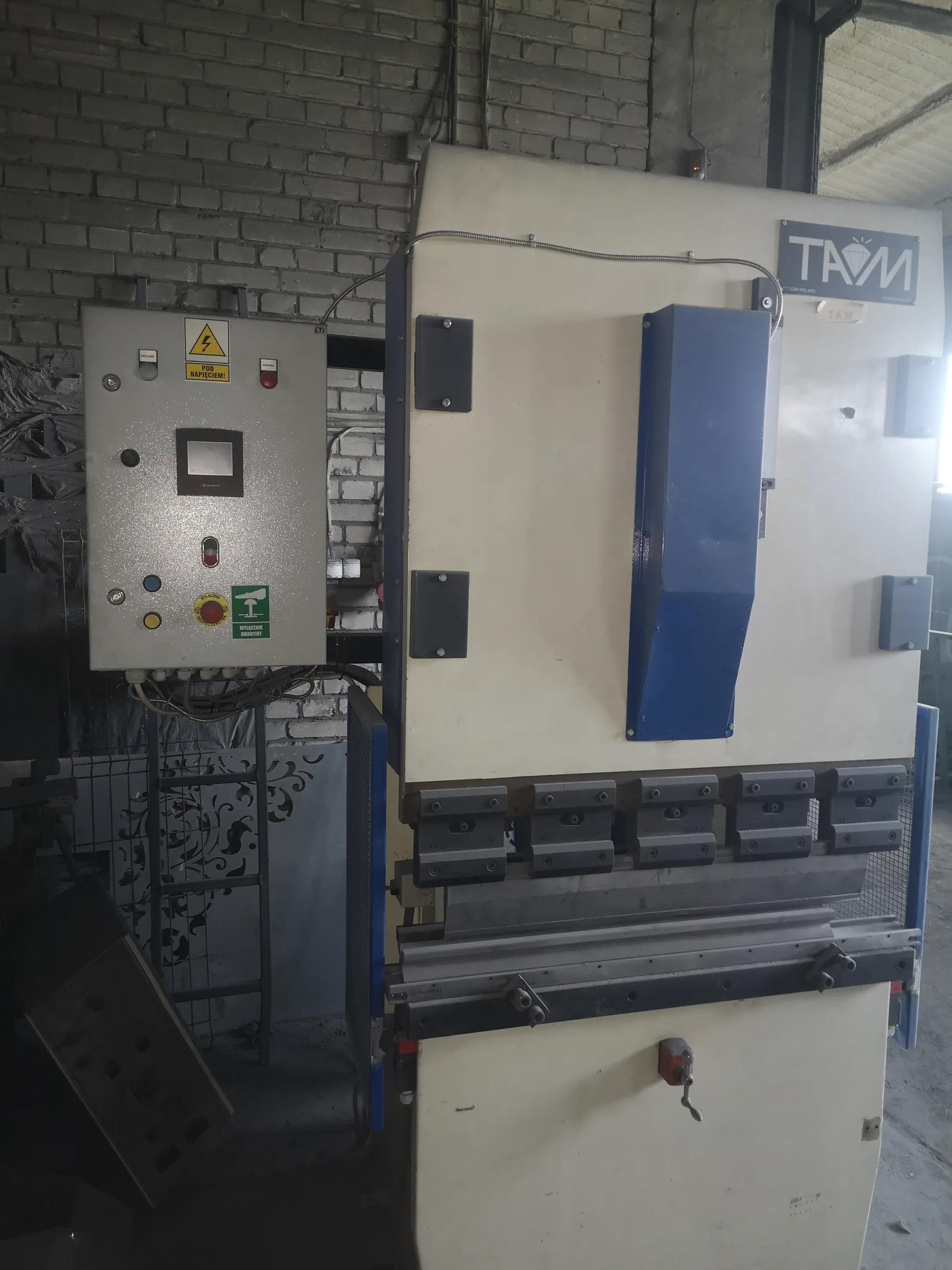 Przemysłowa maszyna o białym i niebieskim kolorze z panelami sterowniczymi. Po lewej stronie szara szafka z przyciskami, czerwonym przyciskiem awaryjnym, żółtym ostrzeżeniem 'POD NAPIĘCIEM!' oraz zielonym znakiem z napisem 'WYŁĄCZNIK AWARYJNY'. Na maszynie widoczny logo 'TAM'. Tło stanowi ceglana ściana. W dolnej części maszyny umieszczone są metalowe elementy zaciskowe.