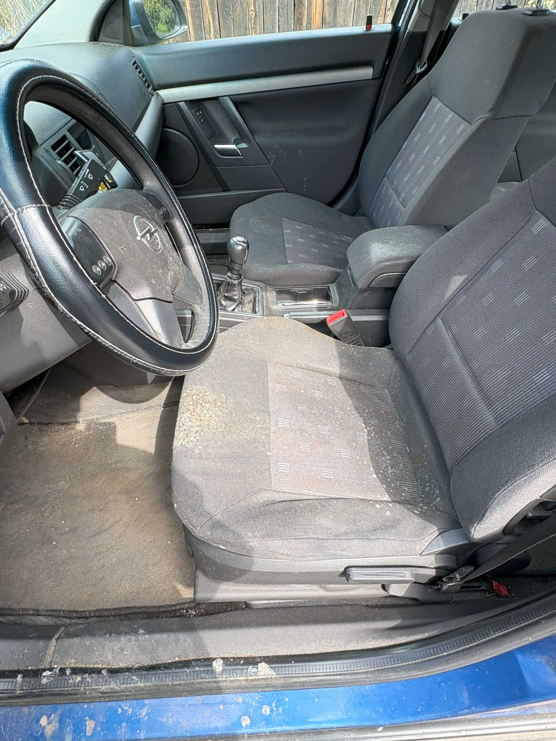 Wnętrze samochodu z widocznym kierownicą z logo, czarnymi siedzeniami w szarym wzorze, brudnymi podłogami i dźwignią zmiany biegów. Drzwi pasażera są otwarte, a na zewnątrz widoczna jest niebieska karoseria. W tle drewniane ogrodzenie. Siedzenia i podłoga pokryte są kurzem i zabrudzeniami.