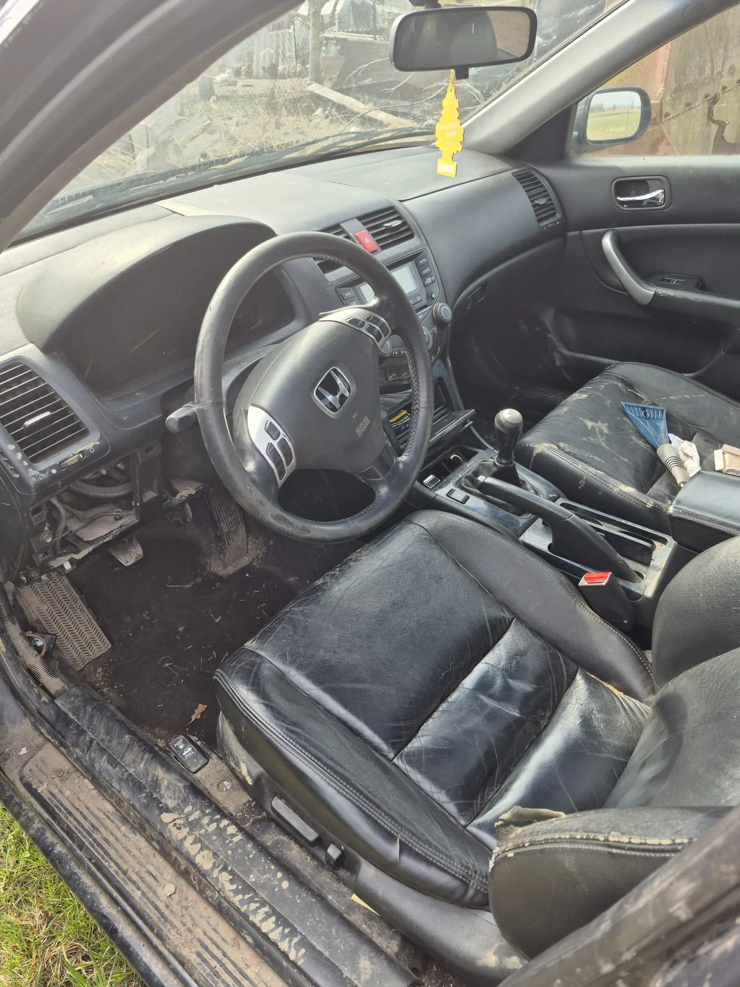 Wnętrze samochodu marki Honda z czarną skórzaną tapicerką w złym stanie. Widoczna kierownica z logo Hondy, deska rozdzielcza, otwarte drzwi kierowcy. Na lusterku wstecznym wiszący żółty odświeżacz powietrza. Wnętrze zabrudzone, na siedzeniach i podłodze widoczne ślady zużycia i brudu. Poza samochodem widoczne zniszczone obiekty i trawa.