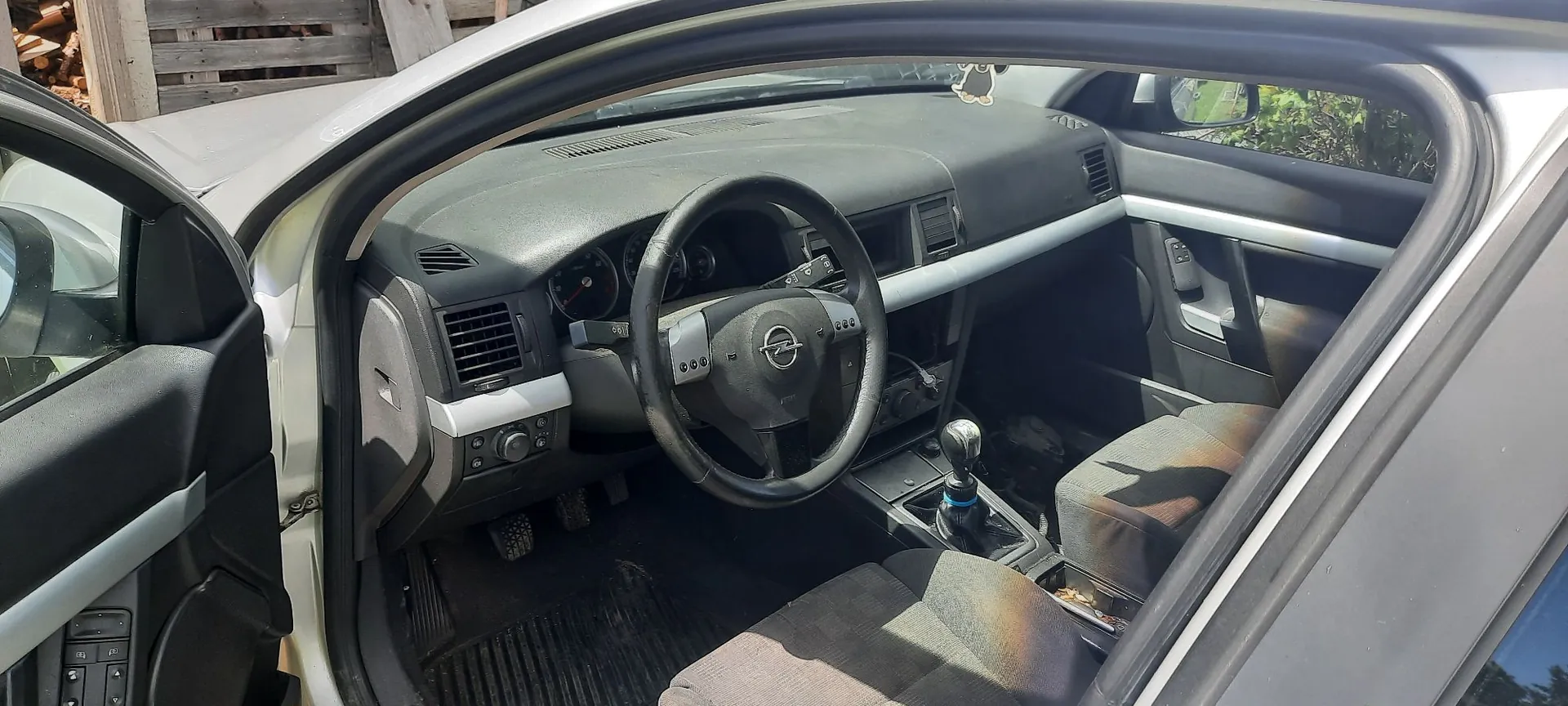 Wnętrze samochodu marki Opel widziane z otwartych drzwi kierowcy. Widoczna jest czarna kierownica z logo producenta, deska rozdzielcza, panel z nawiewami, lewarek zmiany biegów oraz dwa przednie fotele z ciemnej tkaniny. W tle widać fragmenty nadwozia w kolorze białym.