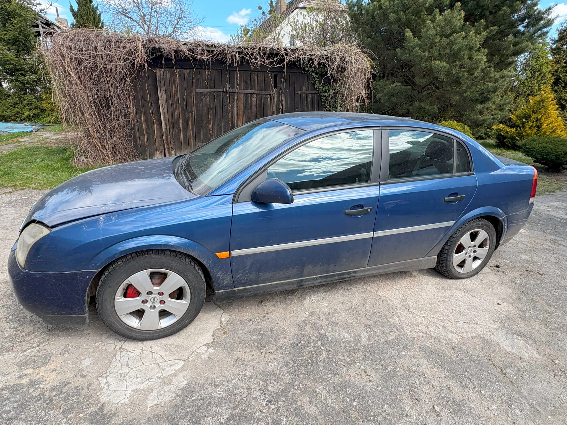 Niebieski samochód osobowy typu sedan zaparkowany na betonowej powierzchni. Samochód ma uszkodzony lewy reflektor przedni, srebrne felgi z czerwonymi zaciskami hamulcowymi oraz pomarańczowy odblask na przednim błotniku. W tle drewniana szopa porośnięta suchymi pnączami, drzewa, krzewy oraz budynek mieszkalny z białą elewacją. Niebo jest błękitne z kilkoma chmurami. Powierzchnia pod samochodem pokryta pęknięciami.