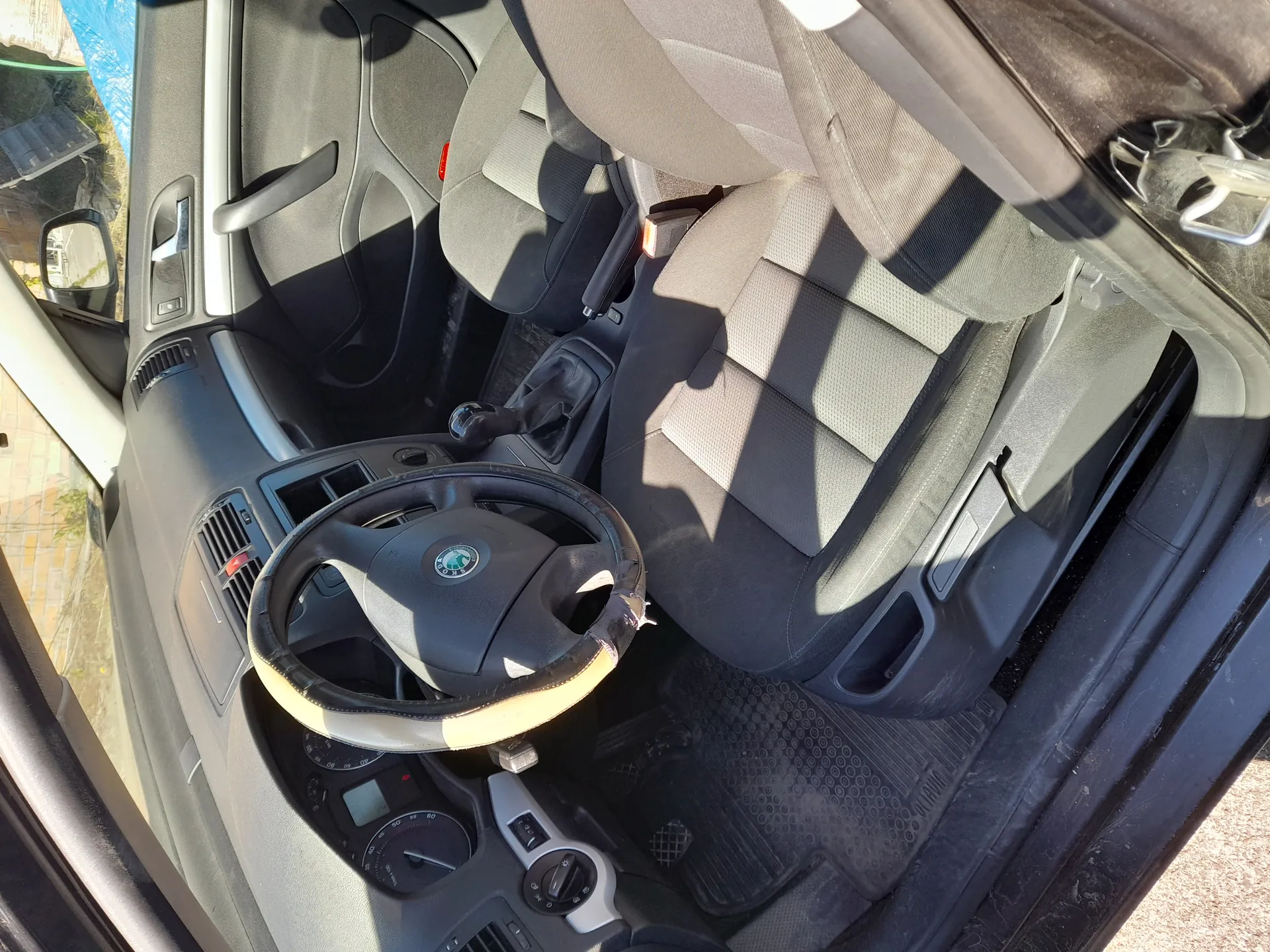 Wnętrze samochodu z widoczną kierownicą z logo Škoda, deską rozdzielczą z wskaźnikami, dźwignią zmiany biegów, siedzeniami tapicerowanymi szarym materiałem, pasami bezpieczeństwa oraz czarnymi dywanikami podłogowymi. Drzwi samochodu są otwarte, a na zewnątrz widoczny jest bruk i fragment zieleni. Światło słoneczne rzuca cienie na wnętrze.