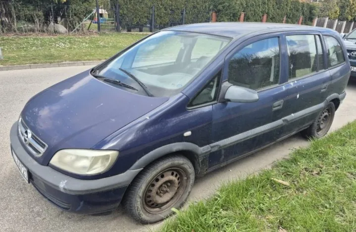 Ciemnoniebieski samochód osobowy marki Opel, zaparkowany na poboczu drogi. Pojazd ma brakującą przednią lewą oponę, widoczne zabrudzenia na karoserii. W tle trawa, ogrodzenie, drzewa oraz elementy placu zabaw. Tablica rejestracyjna częściowo widoczna.