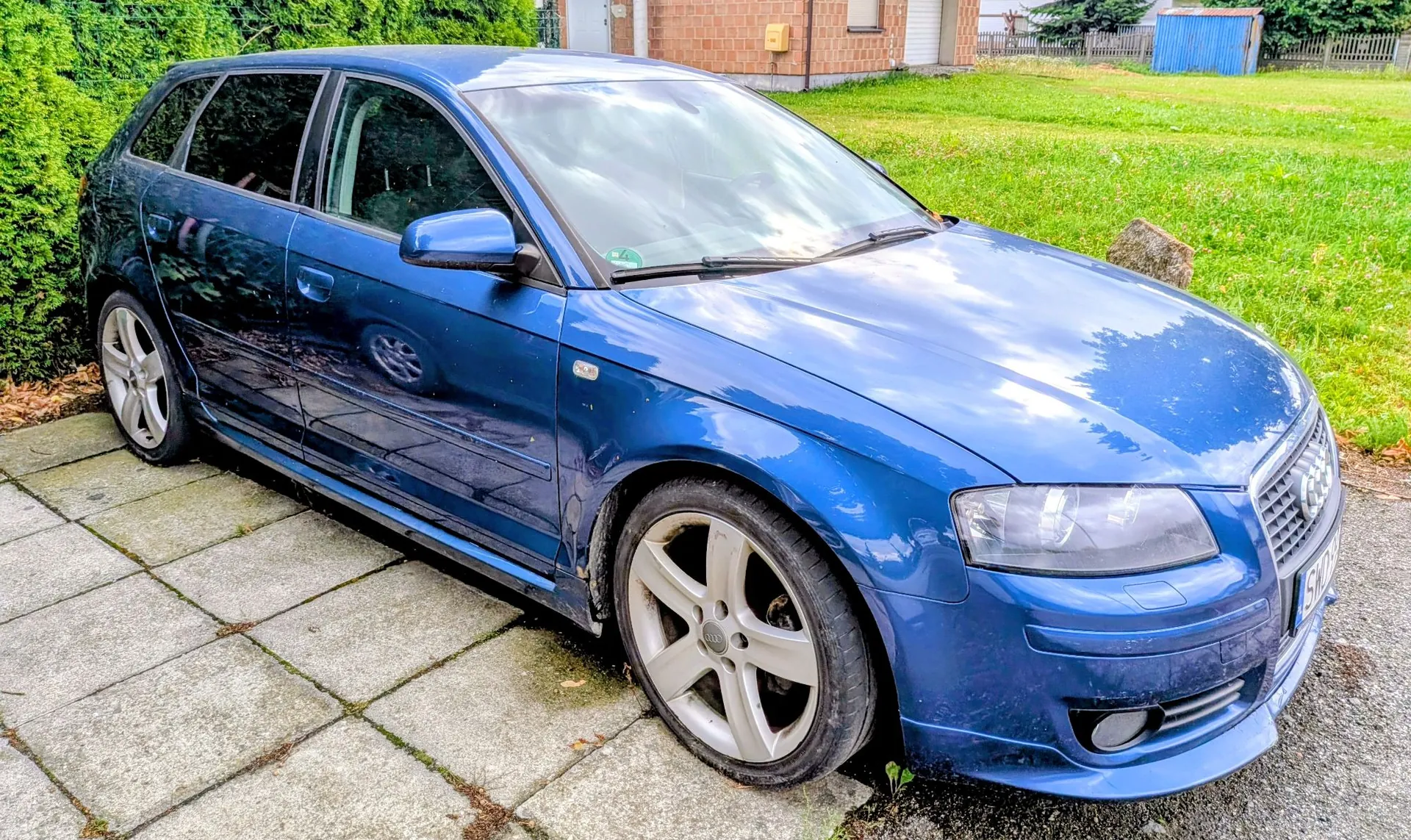 Niebieski samochód marki Audi w nadwoziu kombi, zaparkowany na szarym chodniku z płyt betonowych. Auto jest ustawione pod kątem, widoczny jest przód oraz lewy bok pojazdu. W tle znajduje się zielony trawnik, krzewy oraz fragmenty budynków mieszkalnych.