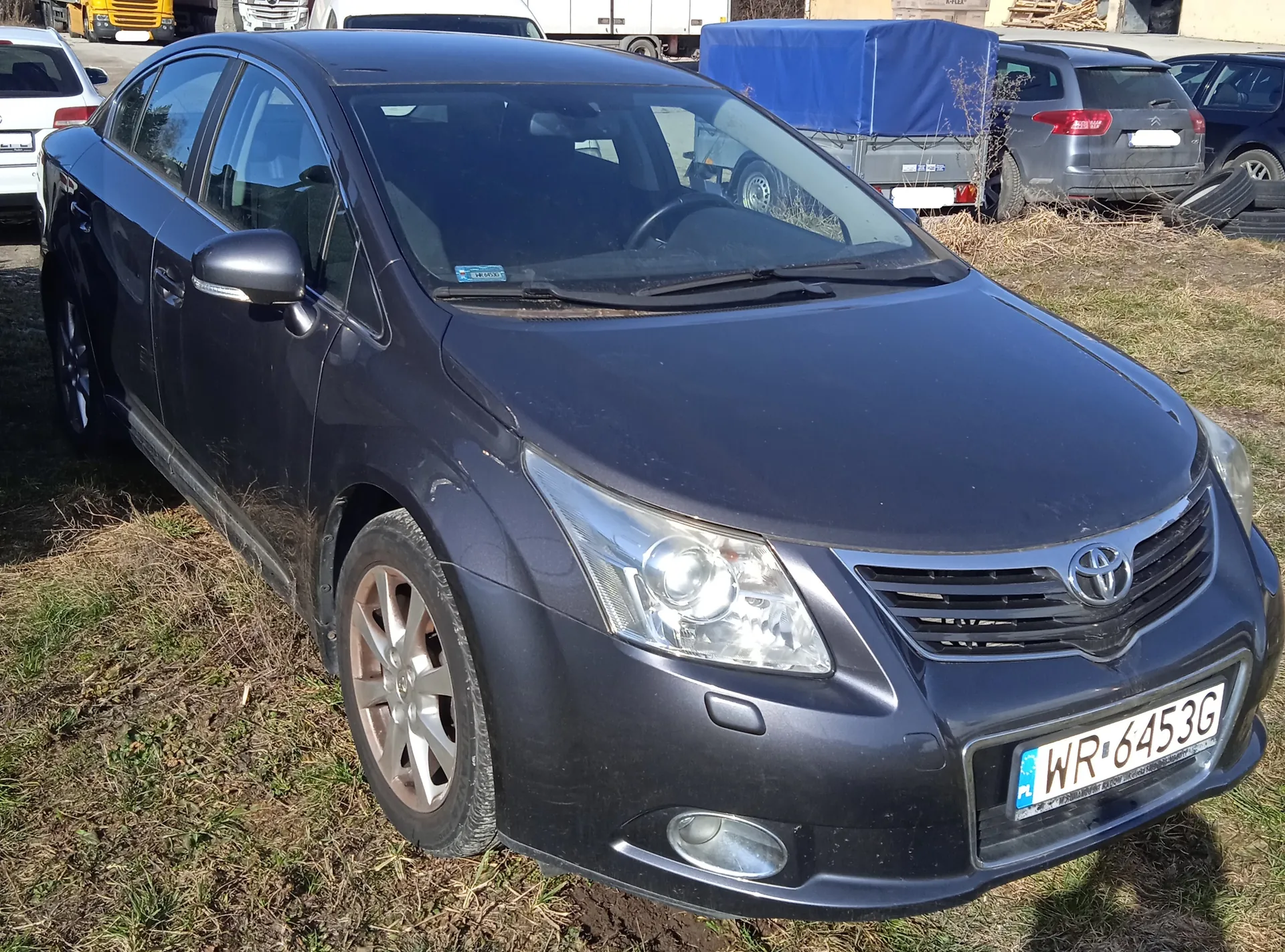 Ciemnoniebieski samochód osobowy marki Toyota z tablicą rejestracyjną WR 6453G (PL) zaparkowany na trawie. Na przedniej szybie widoczna niebieska naklejka. W tle inne pojazdy: biały samochód, szary SUV, ciężarówka z niebieską pokrywą oraz stos opon. Trawa częściowo sucha, na dalszym planie budynki i pojazdy.
