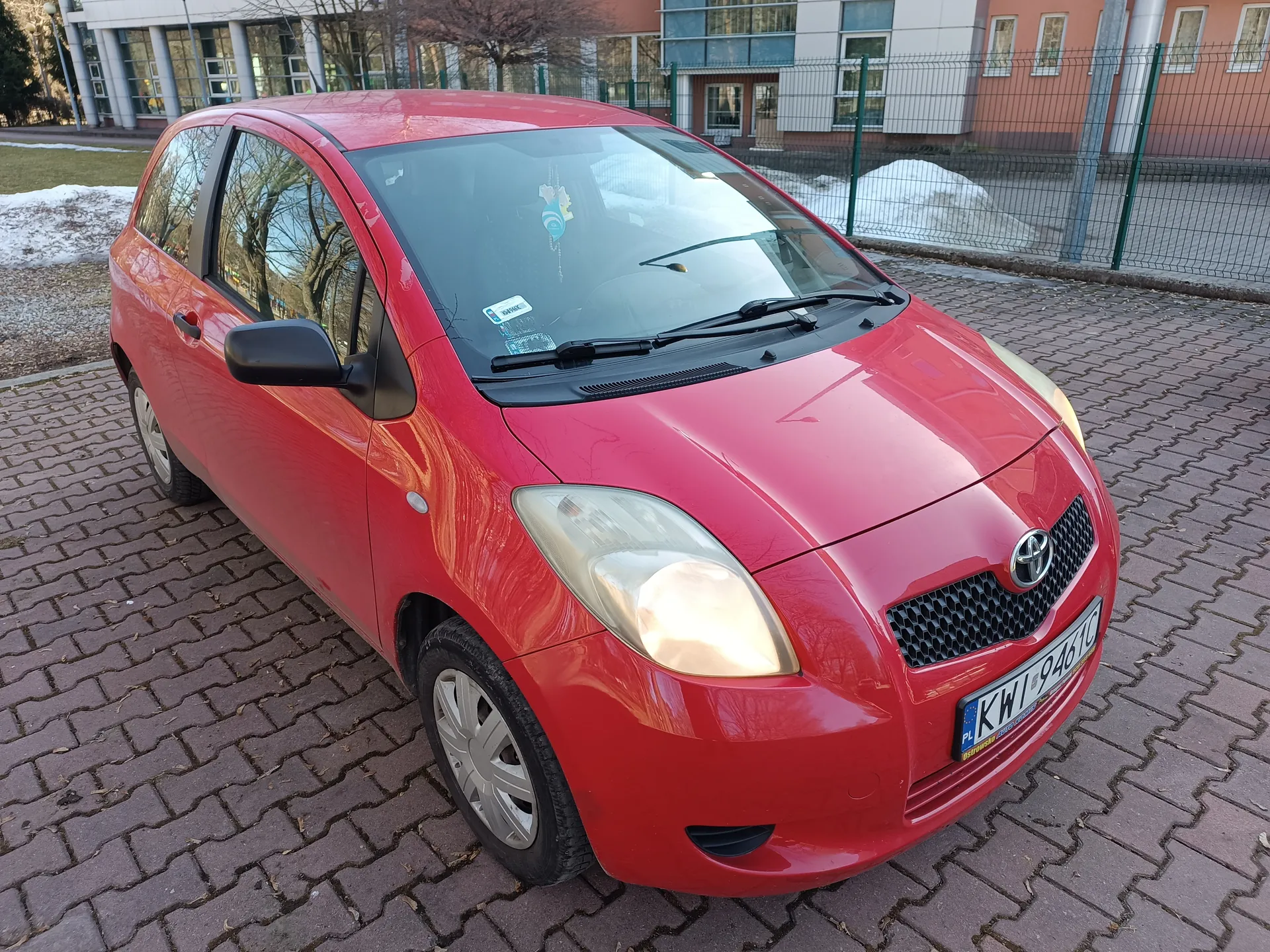 Czerwony samochód marki Toyota z tablicą rejestracyjną KWI 9461C zaparkowany na brukowanej powierzchni. W tle budynki, ogrodzenie, resztki śniegu i drzewa bez liści. Na przedniej szybie widoczne naklejki.
