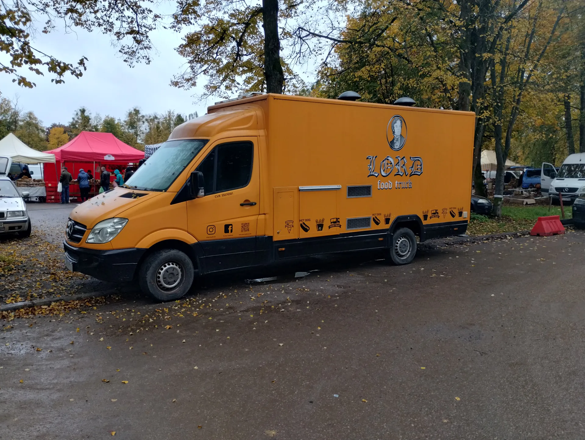 Żółty food truck marki Mercedes-Benz z napisem "LORD food truck" na boku, wraz z ikonami jedzenia (hot dog, pizza) i portretem w okręgu. Pojazd stoi na nawierzchni z rozsypanymi liśćmi, w tle czerwone i białe namioty, ludzie, drzewa z żółtymi liśćmi oraz inne samochody. Niebo jest zachmurzone.