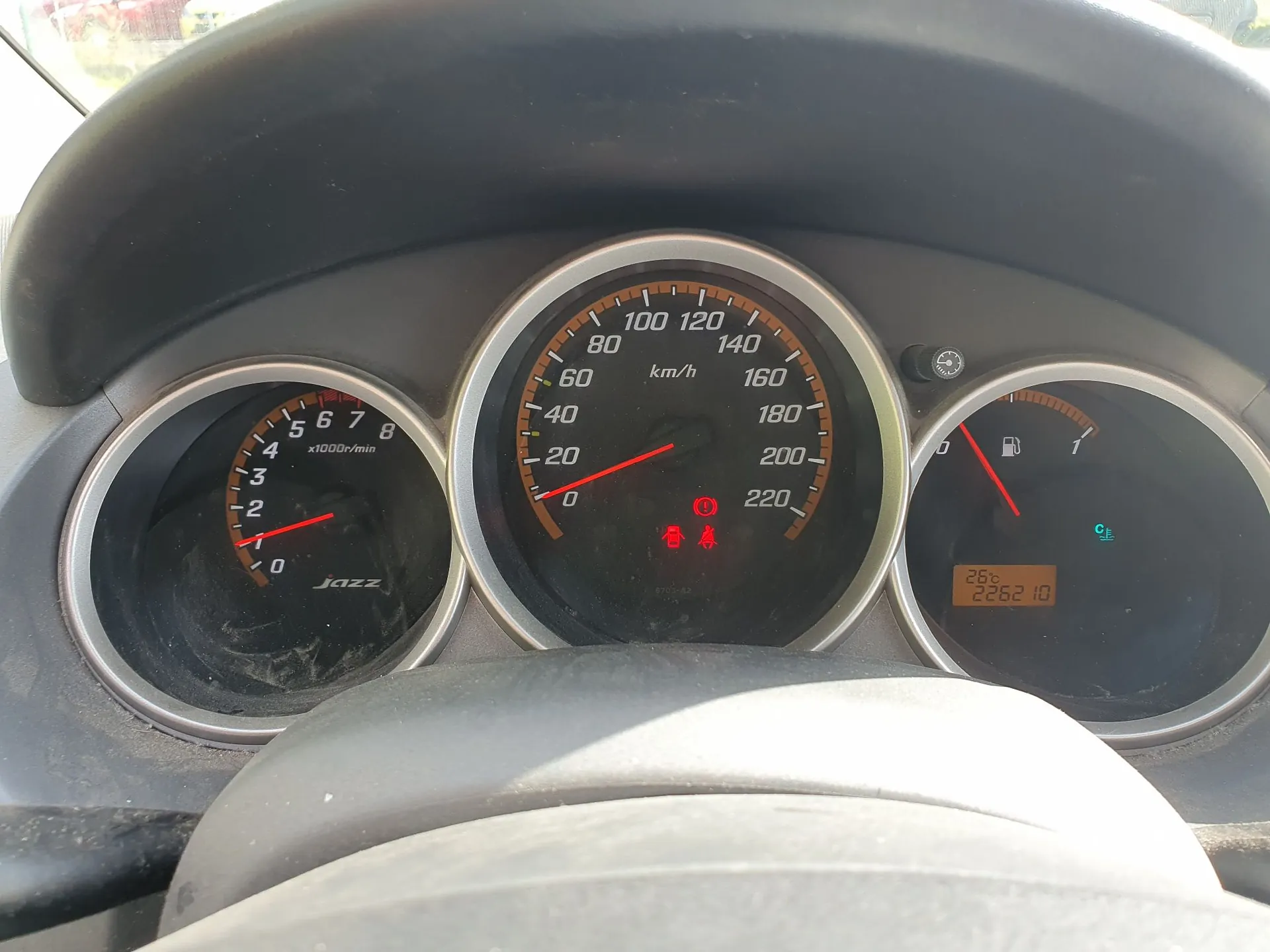 Panel instrumentów samochodu marki Honda Jazz. Widoczne są trzy okrągłe wskaźniki: po lewej obrotomierz z napisem 'x1000r/min' i logo 'Jazz', w centrum prędkościomierz z jednostką 'km/h' i skalą do 220, po prawej wskaźnik poziomu paliwa oraz mały cyfrowy wyświetlacz pokazujący temperaturę '26°C' i przebieg '226210'. Na prędkościomierzu świecą się czerwone kontrolki ostrzegawcze.