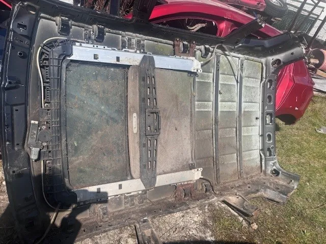 Zdjęcie przedstawia uszkodzoną tylną część nadwozia samochodu, prawdopodobnie bagażnik, leżącą na ziemi. Widoczne są metalowe panele z rdzą, elementy plastikowe oraz przewody elektryczne. W tle znajduje się czerwony samochód z otwartym bagażnikiem oraz trawa i betonowa powierzchnia. Brak czytelnego tekstu.