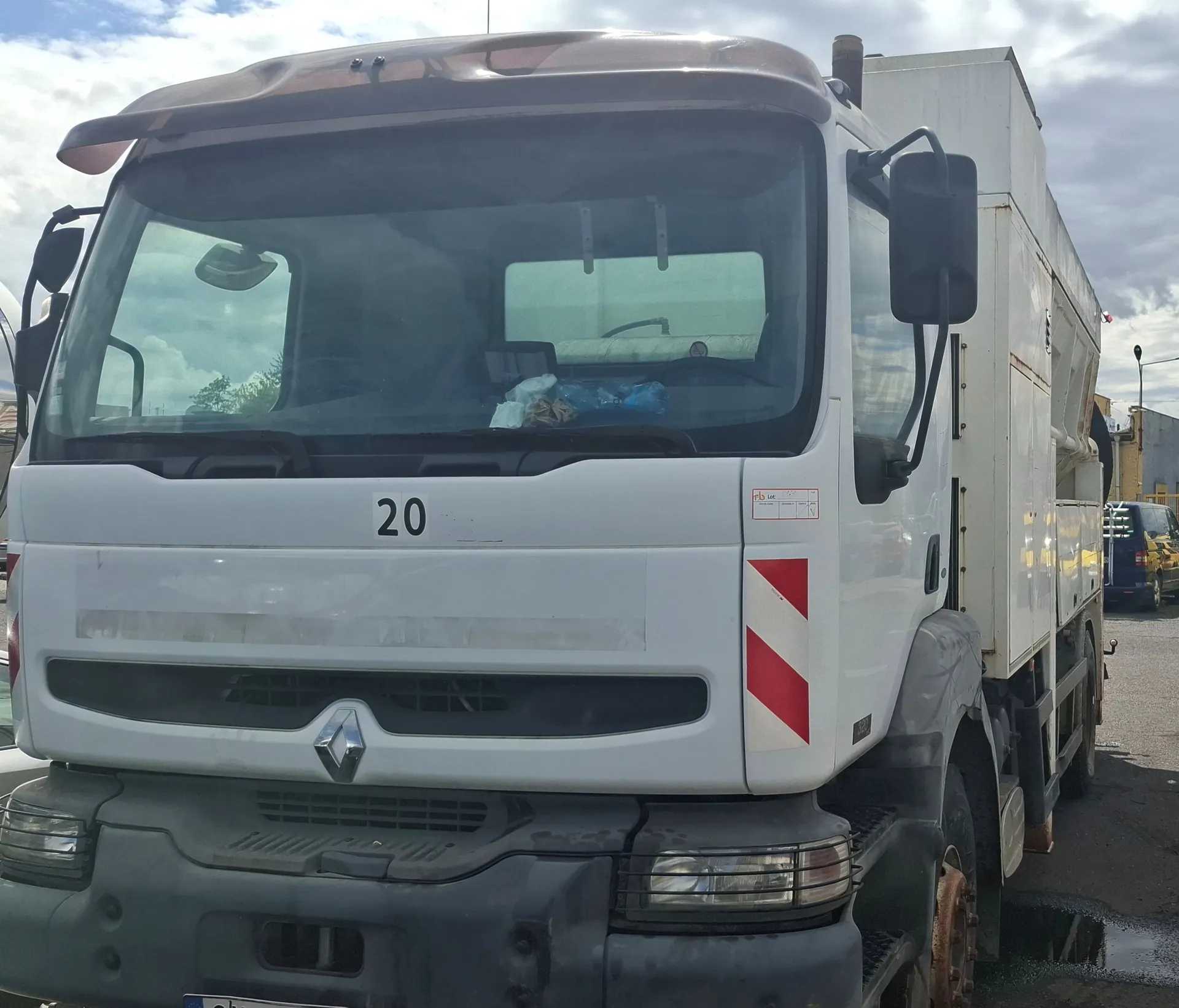 Widok z przodu białej ciężarówki marki Renault. Na masce pojazdu widnieje numer 20, a po prawej stronie znajduje się czerwono-biały pas ostrzegawczy w paski. Pojazd posiada szary zderzak i jest wyposażony w zabudowę typu kontener z tyłu.