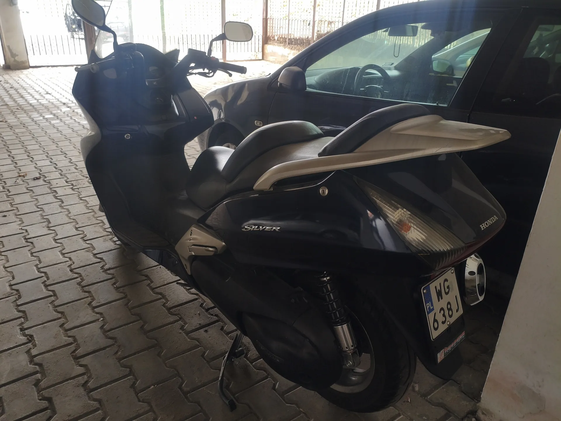 Czarny skuter marki Honda z napisem 'Silver' na boku, z tablicą rejestracyjną PL WG 638J. Skuter stoi na brukowanej powierzchni, obok niego ciemny samochód osobowy. W tle metalowe ogrodzenie i fragment budynku. Oba pojazdy są zaparkowane, a tablica rejestracyjna skutera jest czytelna i poziomo ustawiona.
