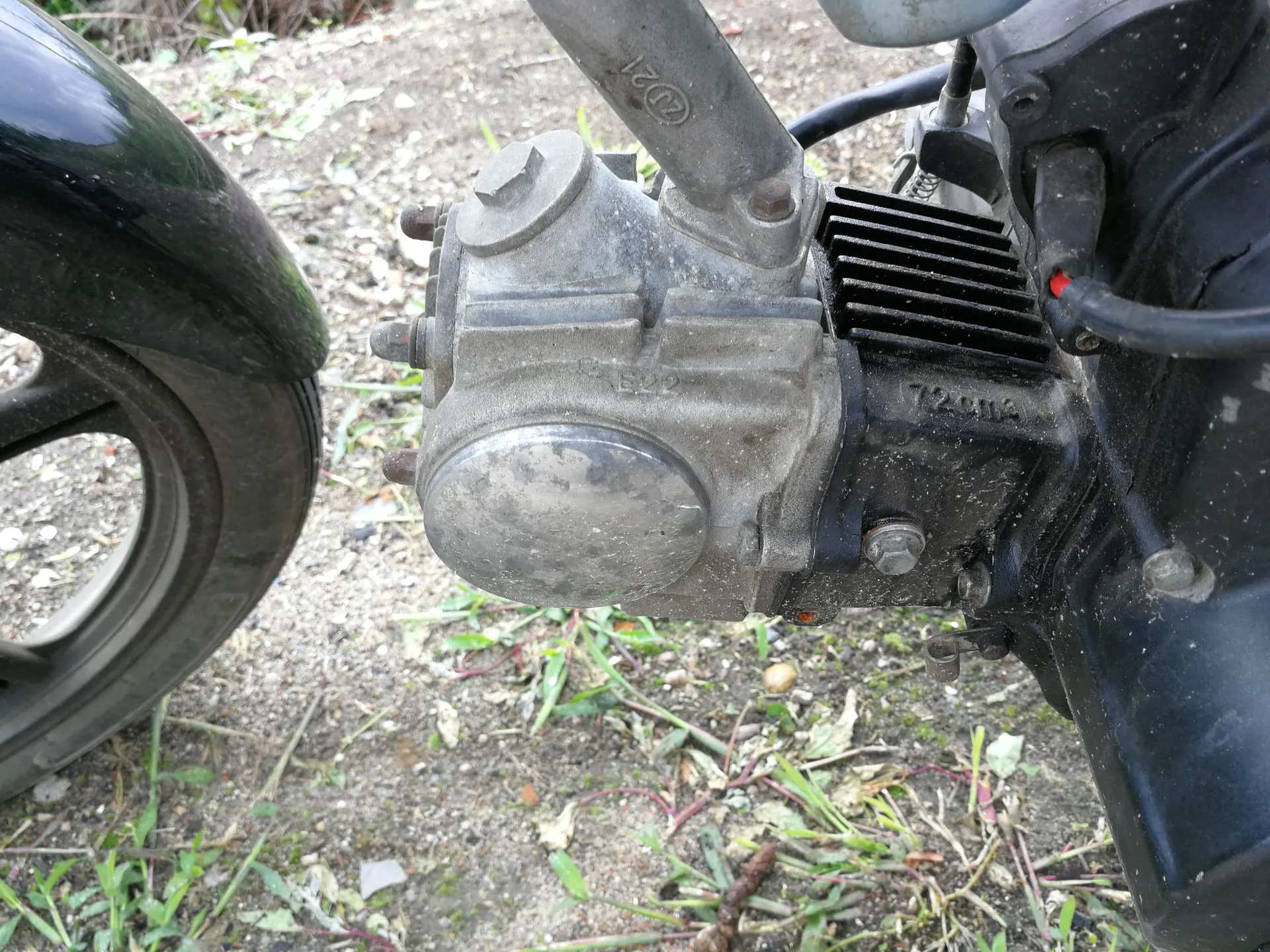 Zbliżenie na silnik motocykla lub skutera. Widoczna jest metalowa obudowa silnika z żebrowanym systemem chłodzenia, śrubami montażowymi i okrągłą osłoną. Po lewej stronie znajduje się fragment czarnego błotnika i opony. Tło stanowi ziemia z kępkami trawy.