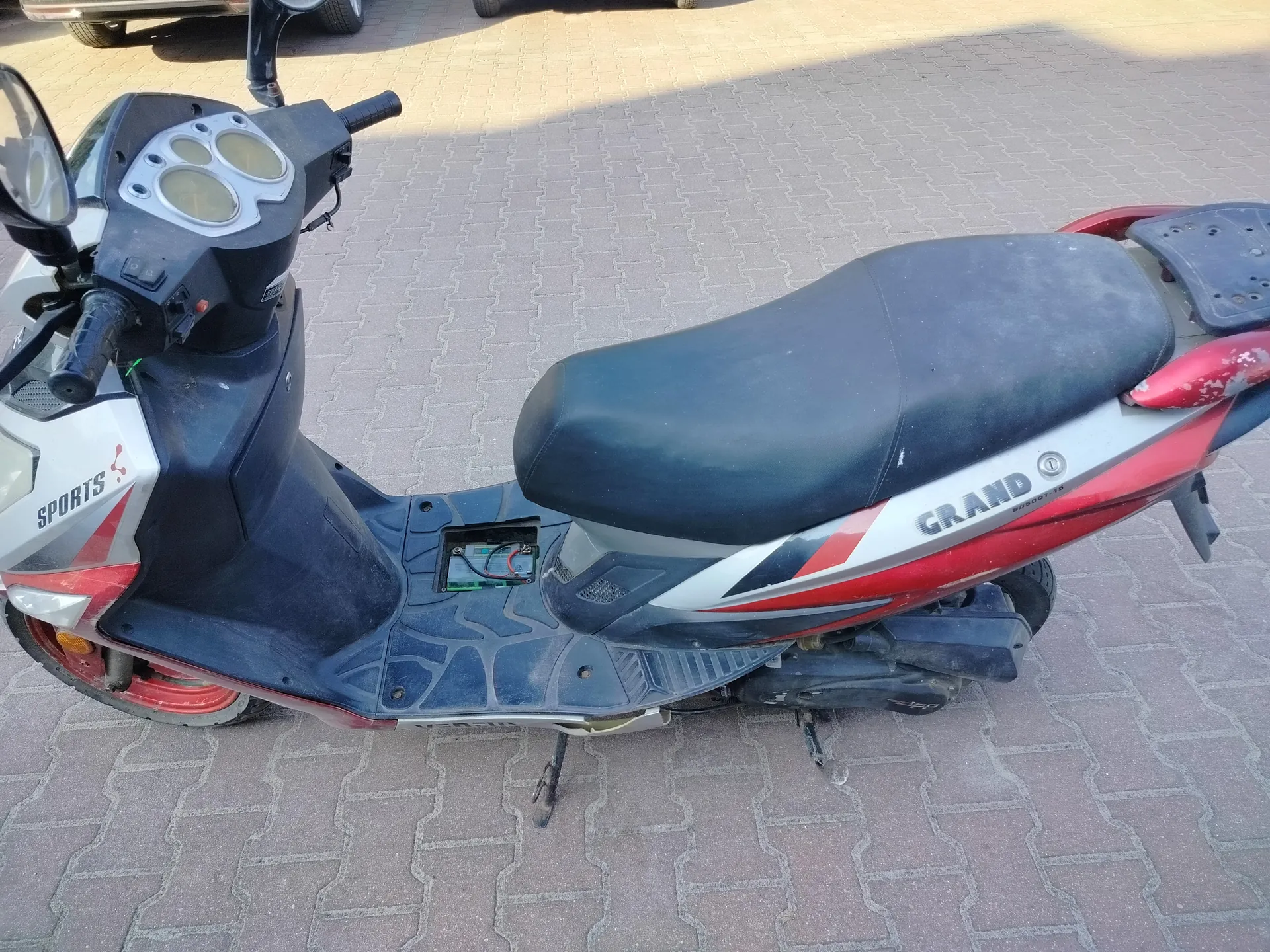 Scooter w kolorach czerwonym, czarnym i srebrnym zaparkowany na chodniku z kostki brukowej. Na przedniej części widoczny napis SPORTS, a na boku napis GRAND. Czarna siedziba, kierownica z lusterkami, trzy okrągłe wskaźniki na desce rozdzielczej. Pod siedziskiem odkryty komponent elektroniczny z przewodami. W tle fragment koła innego pojazdu.