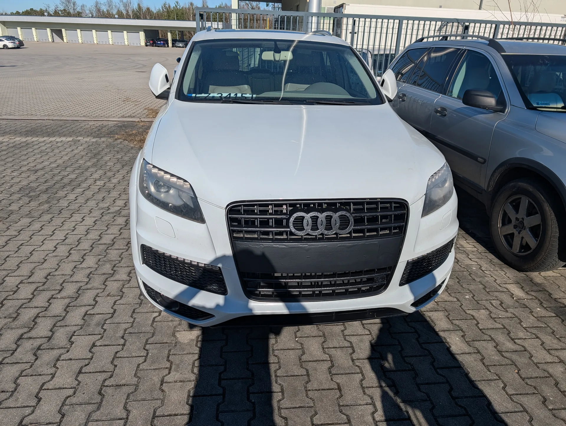 Biały samochód Audi typu SUV z widoczną tablicą rejestracyjną 73415 zaparkowany na brukowanym placu parkingowym. Obok niego stoi szary samochód SUV. W tle budynki, metalowe ogrodzenie oraz inne zaparkowane pojazdy. Jasne niebo, słoneczny dzień.