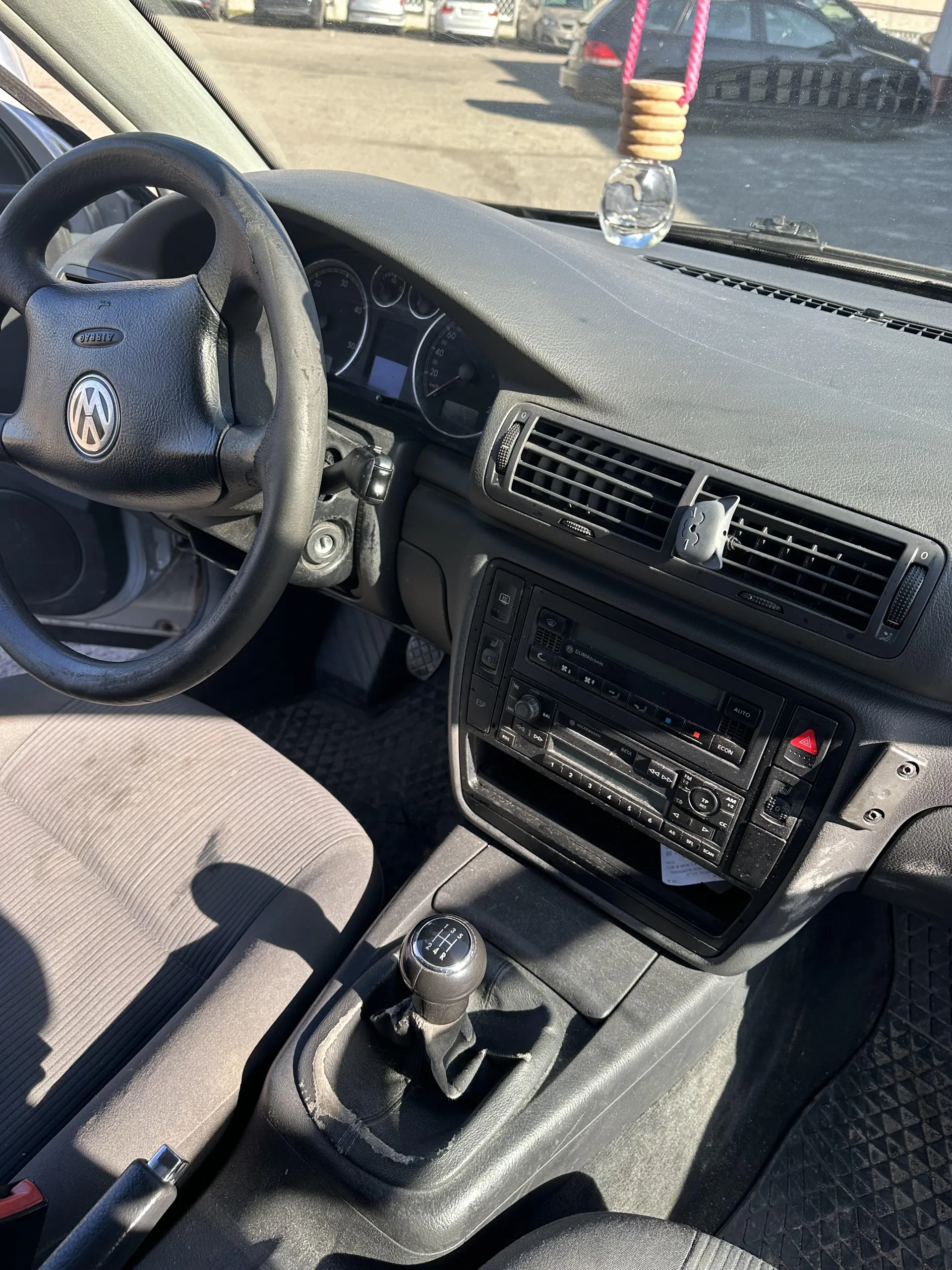 Wnętrze samochodu z widocznym kierownicą z logo i napisem AIRBAG, analogowymi wskaźnikami prędkości i obrotomierzem, manualną skrzynią biegów z oznaczeniami 1-5 i R, centralnym panelem z radiem, przyciskami AUTO, ECON oraz czerwonym przyciskiem alarmu. Na desce rozdzielczej wisząca ozdoba w postaci szklanej butelki z drewnianym elementem i różową sznurówką. Szare siedzenia z pionowymi prążkami, czarne dywaniki z geometrycznym wzorem. Za przednią szybą widoczny parking z zaparkowanymi samochodami i budynkiem w tle.