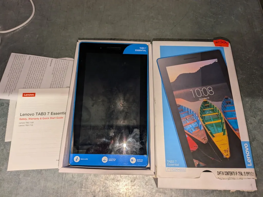 Zestaw produktów marki Lenovo ułożony na szarej powierzchni. Po lewej stronie znajduje się biała instrukcja obsługi z napisem 'Lenovo TAB3 7 Essential Safety, Warranty & Quick Start Guide' oraz modele 'Lenovo TB3-710F' i 'Lenovo TB3-710X'. W środku, w białym pudełku, leży tablet z czarnym ekranem i niebieskimi ramkami, z napisem 'TAB3 7 ESSENTIAL' w prawym górnym rogu. Po prawej stronie znajduje się białe opakowanie kartonowe z grafiką tabletu wyświetlającego kolorową tapetę z łodziami i godziną '10:08', a poniżej napisy 'TAB3 7 Essential' oraz '7" | 1024x600'. W prawym górnym rogu opakowania przyklejona jest czerwona etykieta z ceną '230'.