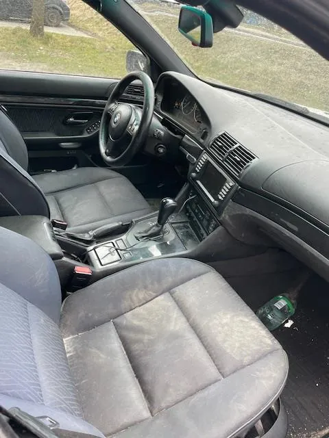 Wnętrze samochodu z czarnymi skórzonymi fotelami, kierownicą z logo, centralną konsolą z przyciskami i dźwignią biegów. Na podłodze pasażera widoczna zielona butelka. Przez przednią szybę widoczny trawnik i drzewo.