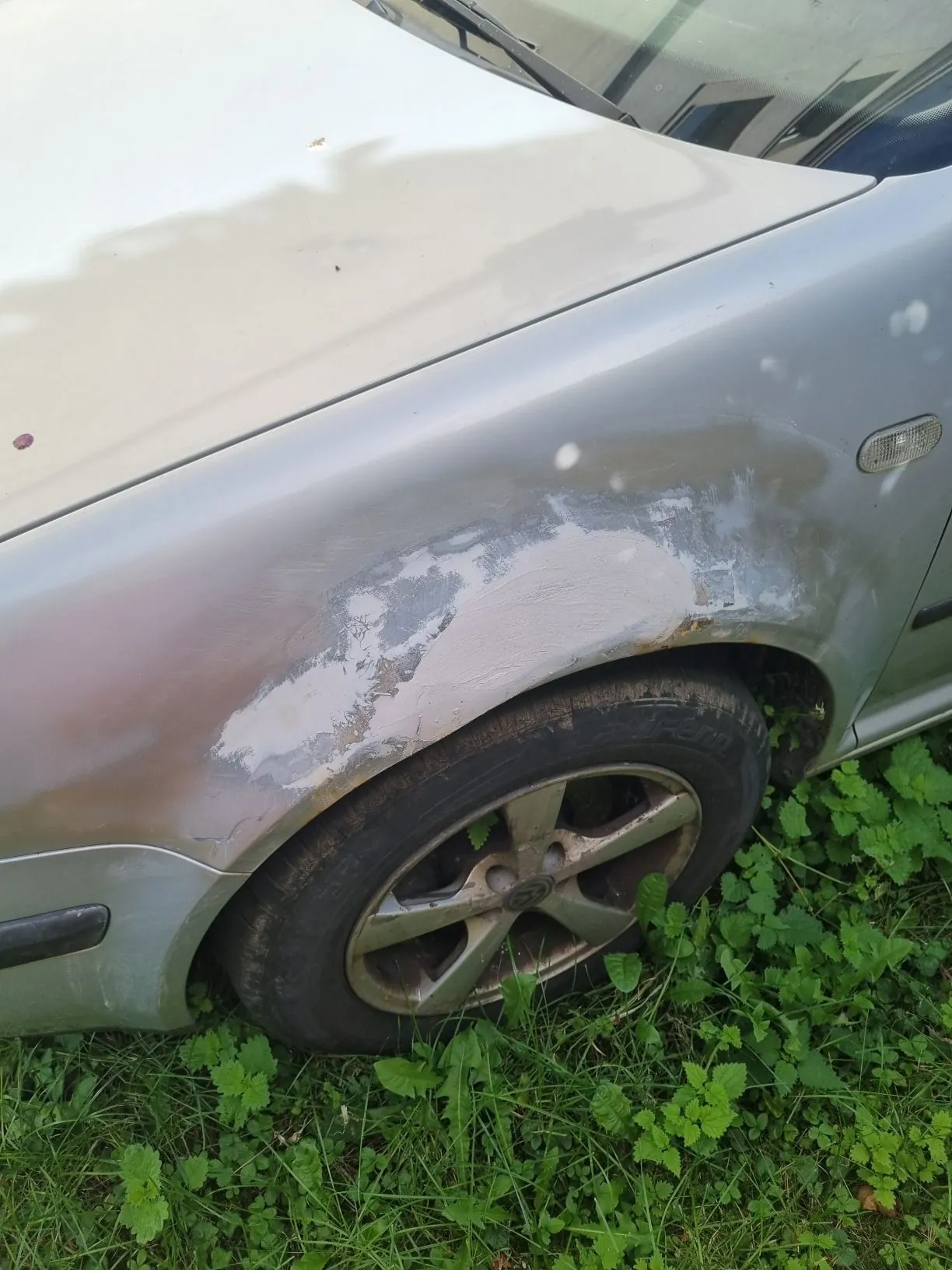 Zbliżenie na przedni prawy błotnik srebrnego samochodu, który jest uszkodzony. Lakier w górnej części nad kołem jest zdrapany, odsłaniając jasnoszarą podkład i miejsca z korozją. Widoczna jest opona z brudną, pięcioramienną felgą aluminiową, zaparkowana na zielonej trawie.