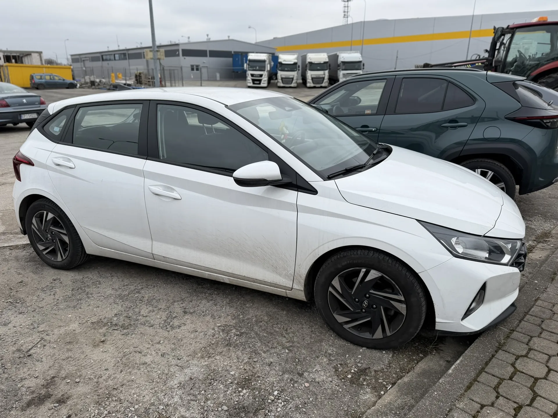 Biały samochód hatchback zaparkowany na żwirowym i brukowanym placu, obok niego ciemnozielony SUV. W tle kilka białych ciężarówek ustawionych przed dużym budynkiem przemysłowym z żółtą listwą. Widoczne są również inne pojazdy, w tym czerwony pojazd budowlany i niebieski samochód. Niebo jest zachmurzone.