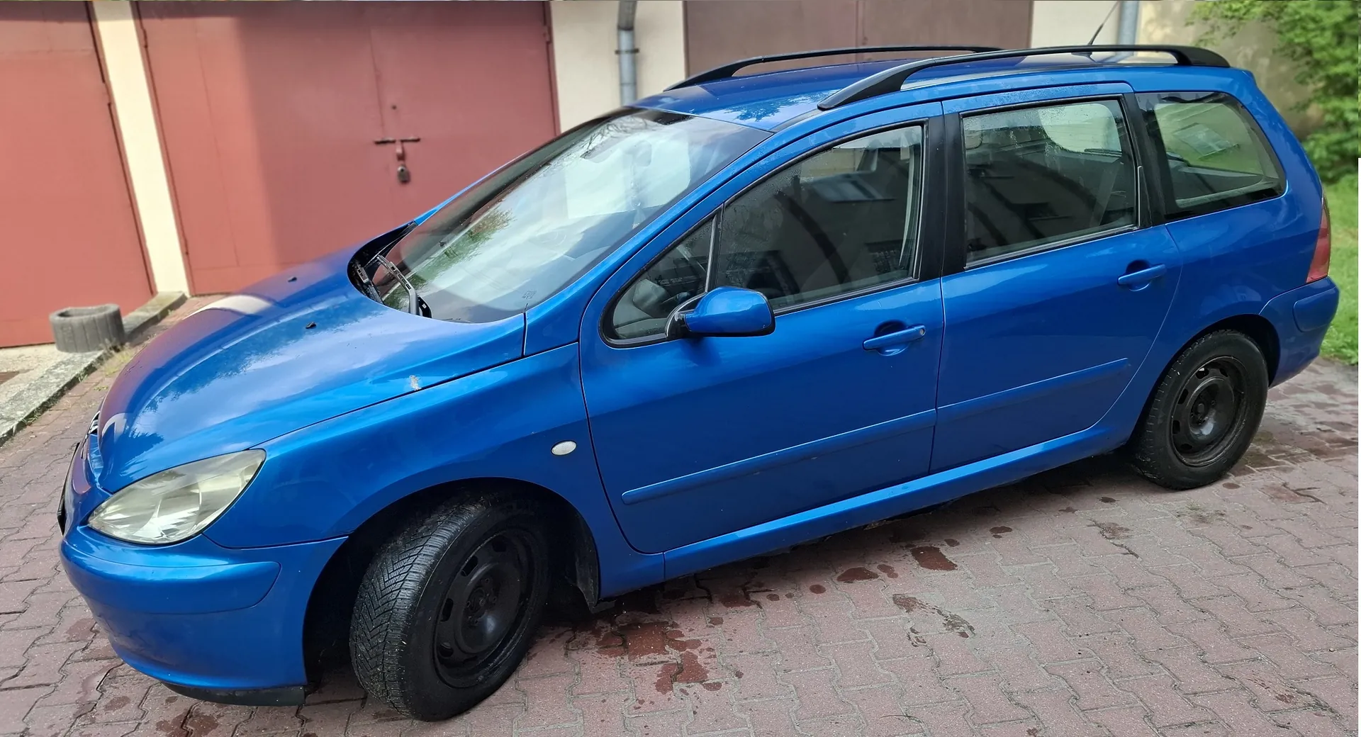 Niebieski samochód typu kombi zaparkowany na kostce brukowej przed budynkiem z czerwonymi drzwiami garażowymi. Pojazd posiada czarne relingi dachowe oraz czarne felgi stalowe.