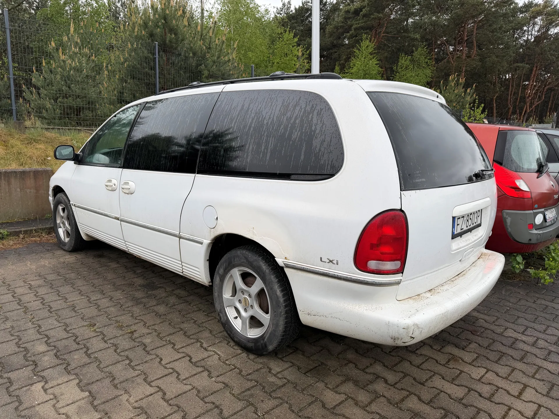 Biały minivan typu kombi z widocznym oznaczeniem "LXi" na tylnej części, z tablicą rejestracyjną FZ 8503P. Pojazd stoi na parkingowym placu z kostki brukowej, obok czerwonego samochodu. W tle siatka ogrodzeniowa, zielone drzewa i krzewy. Szkła minivana są przyciemnione, a nadwozie pokryte jest lekkim brudem. Opony i felgi są widoczne, a na tle widać również fragment innych zaparkowanych pojazdów.