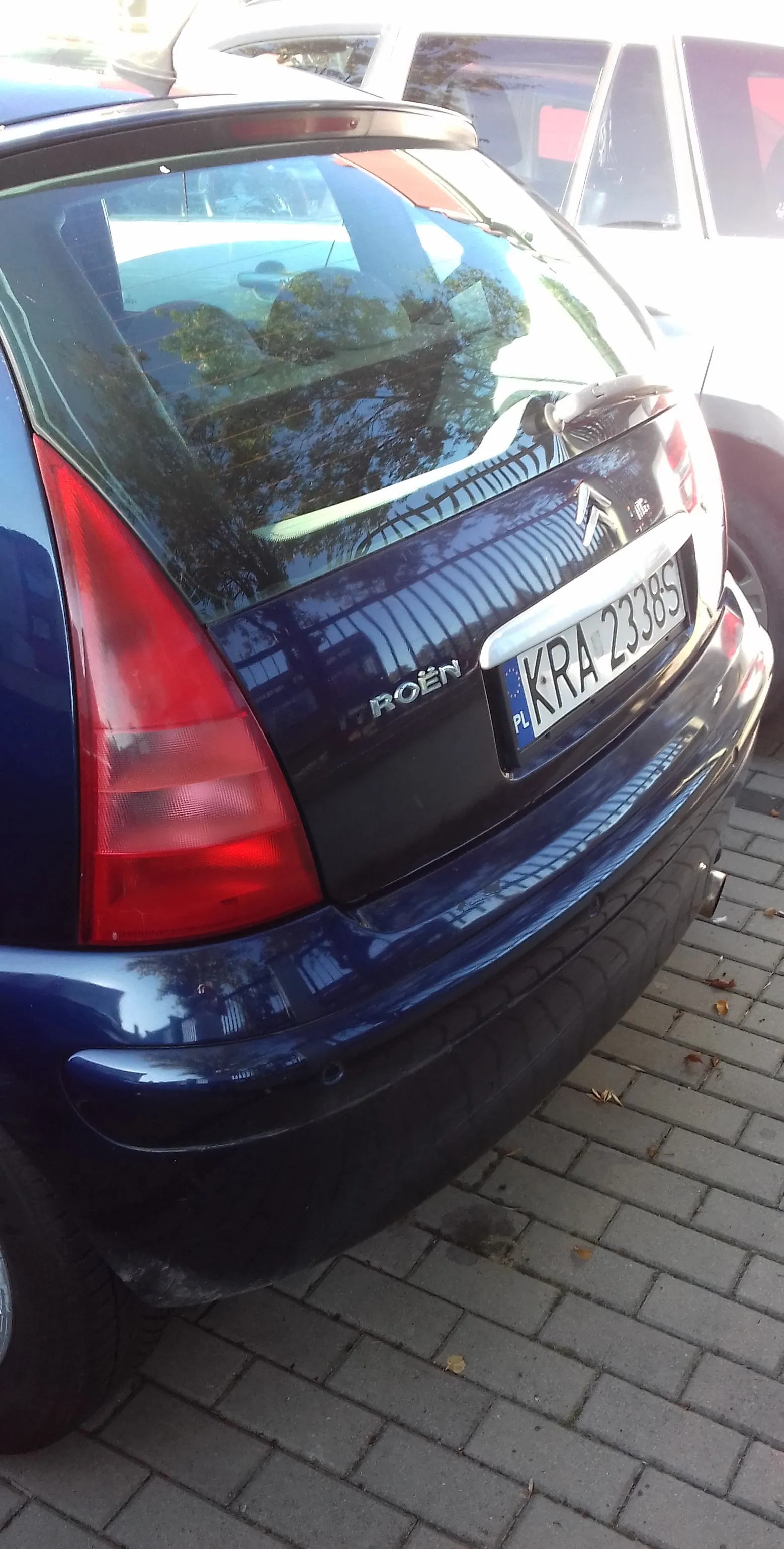 Tylna część ciemnoniebieskiego samochodu marki Citroën. Widoczna jest tylna szyba z odbiciem drzew, czerwona lampa tylna po lewej stronie oraz tablica rejestracyjna o numerze KRA 23385. Samochód zaparkowany jest na kostce brukowej.