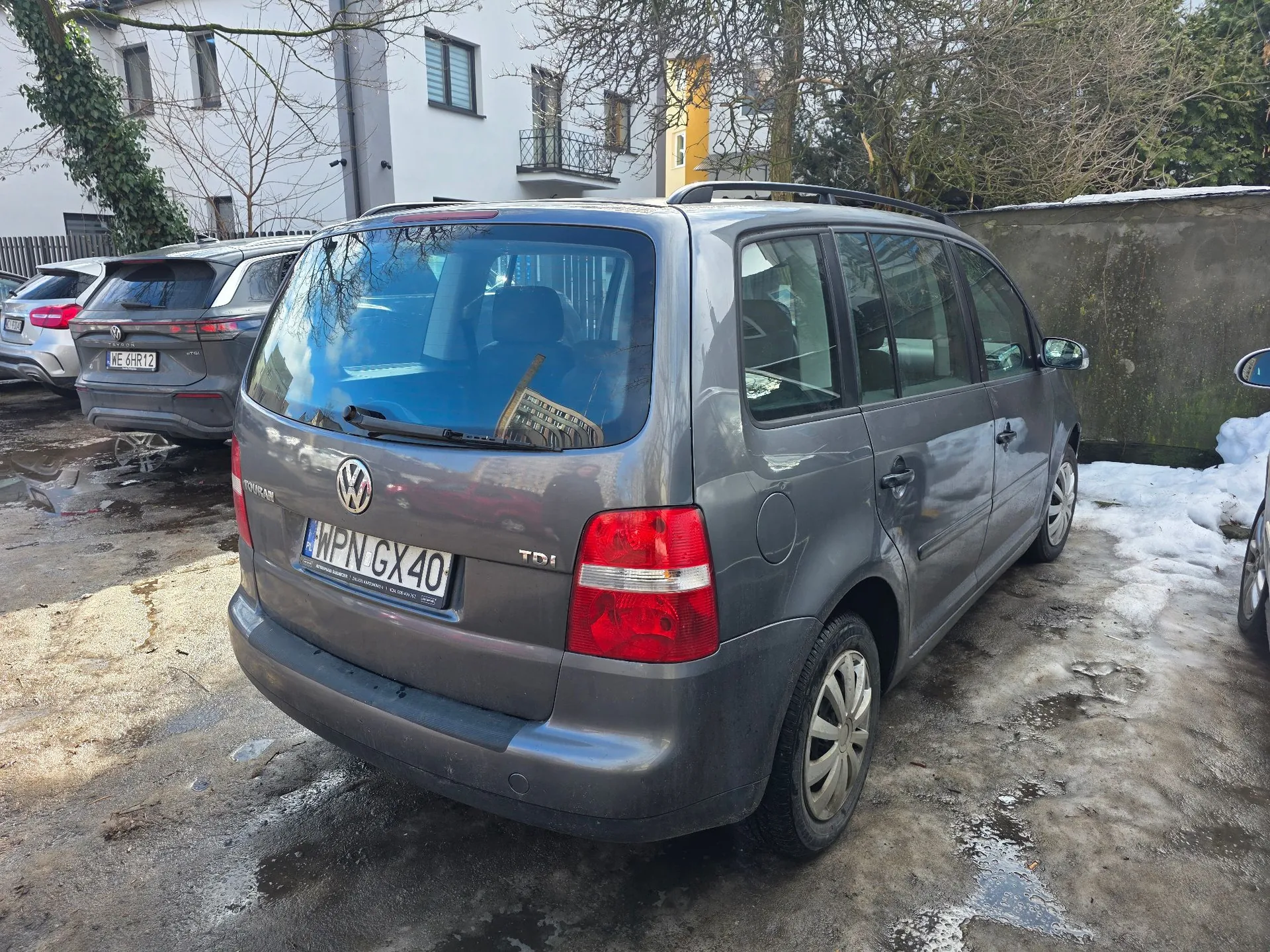 Szary samochód marki Volkswagen Touran w wersji kombi, zaparkowany tyłem do obiektywu na błotnistym podłożu z resztkami śniegu. Samochód ma polską tablicę rejestracyjną o numerze WPN GX40 oraz oznaczenie silnika TDI. W tle widoczny jest biały budynek mieszkalny, drzewa bez liści oraz inny zaparkowany samochód po lewej stronie.