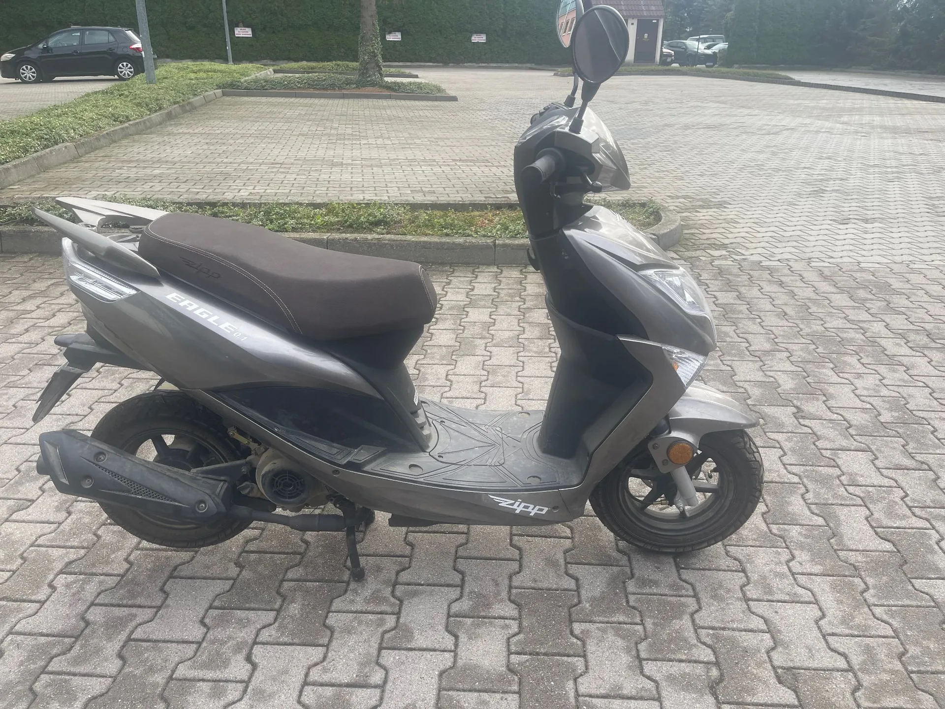 Szary skuter marki Zipp, model Eagle, zaparkowany bokiem na kostce brukowej. Na boku skutera widnieją napisy 'EAGLE' oraz 'zipp'. Pojazd ma czarne felgi i brązowe siedzenie.